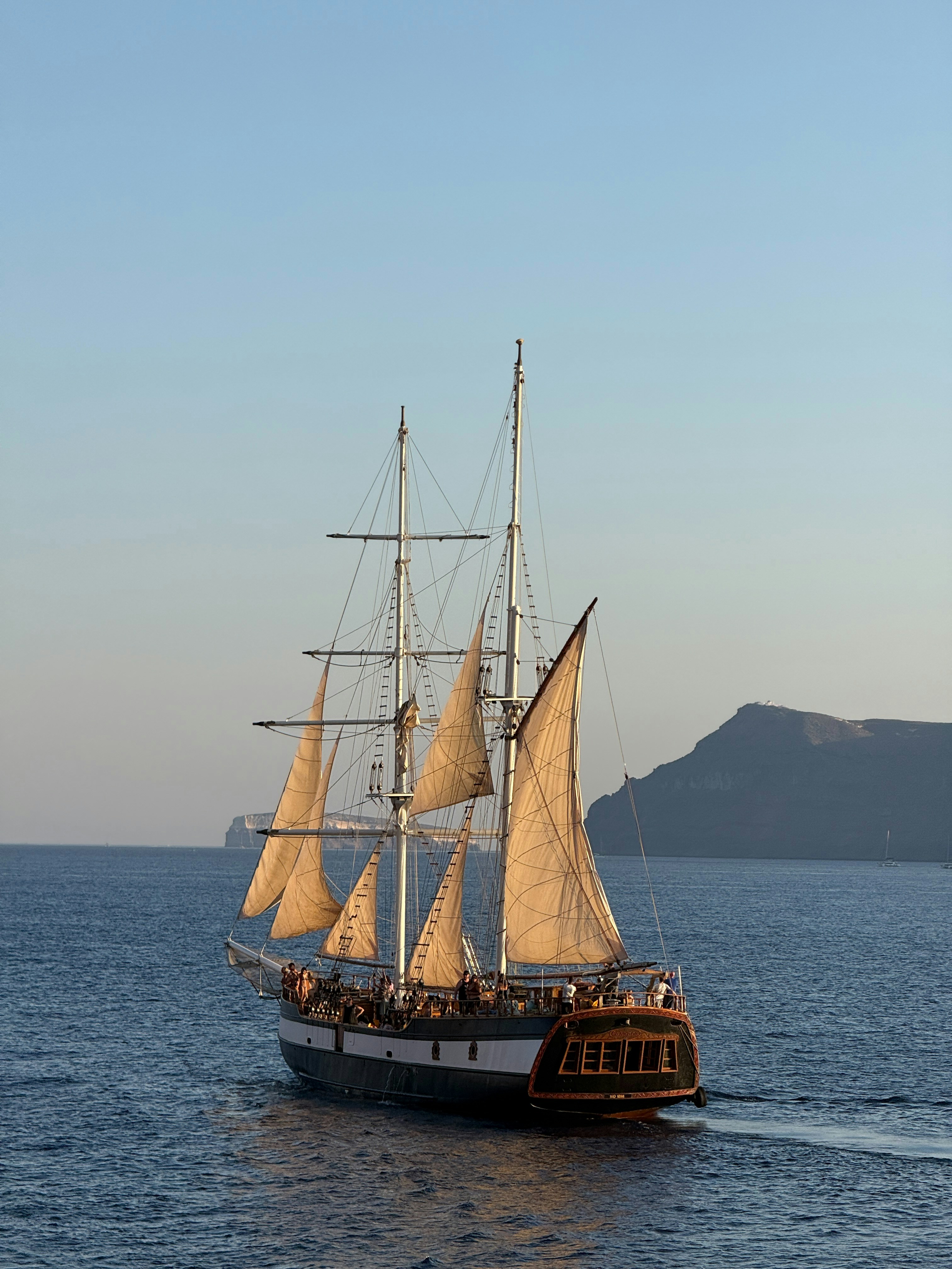 A two-masted sailing ship with tan sails on blue water near rocky cliffs
