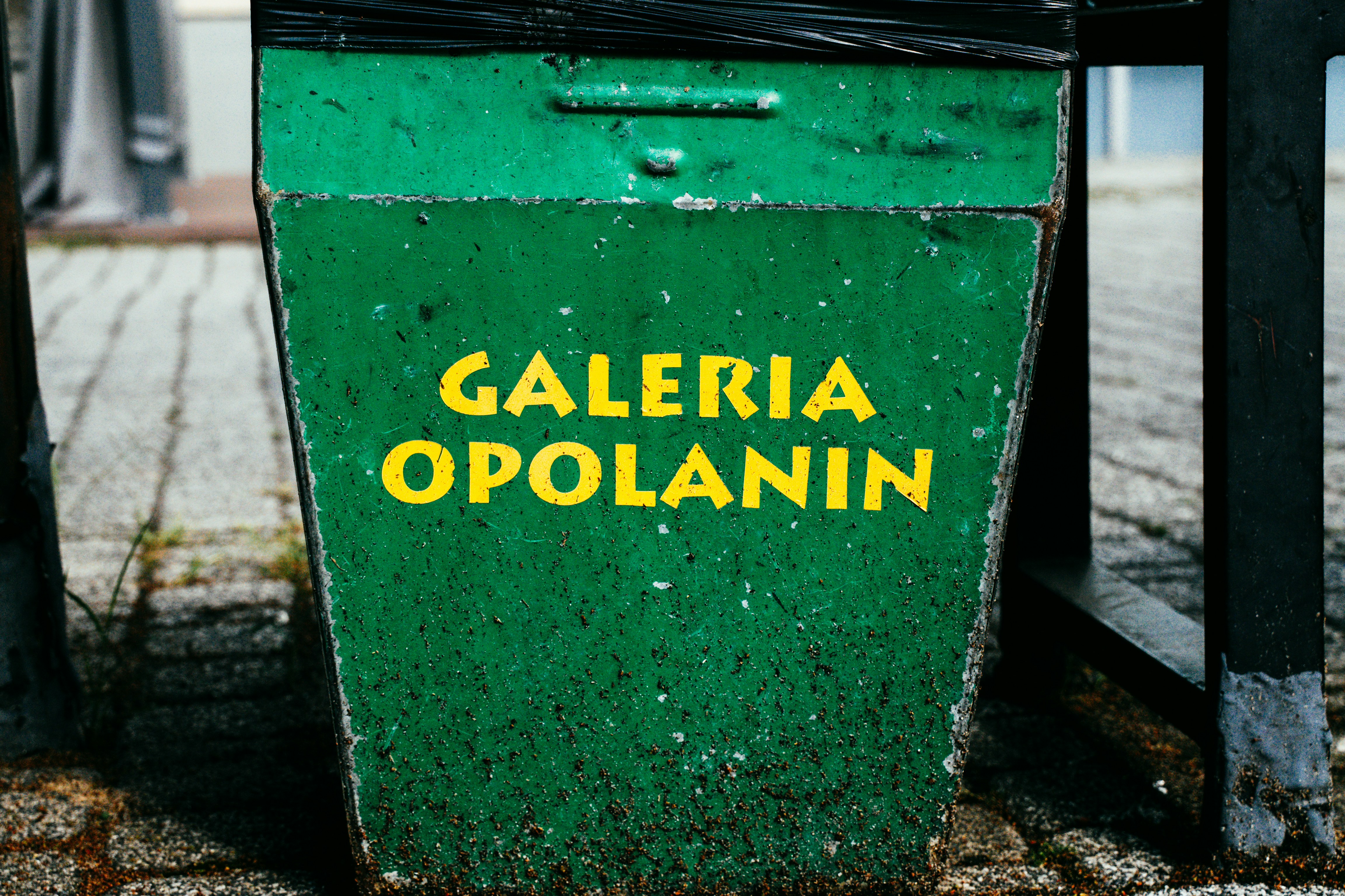 Uma lixeira de metal verde envelhecida com texto amarelo dizendo "GALERIA OPOLANIN"