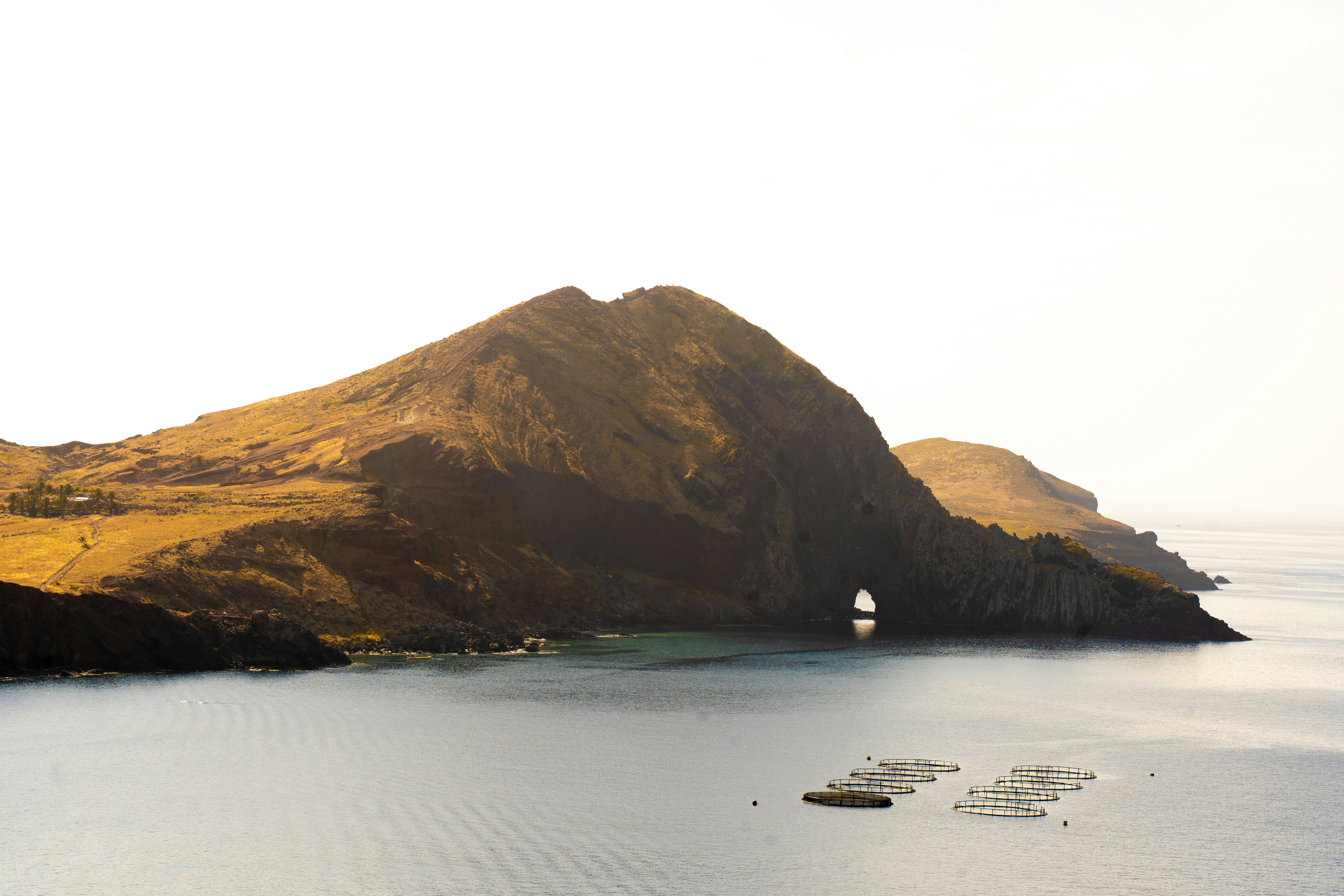A rocky coastal mountain with a natural arch above calm water and fish farms.