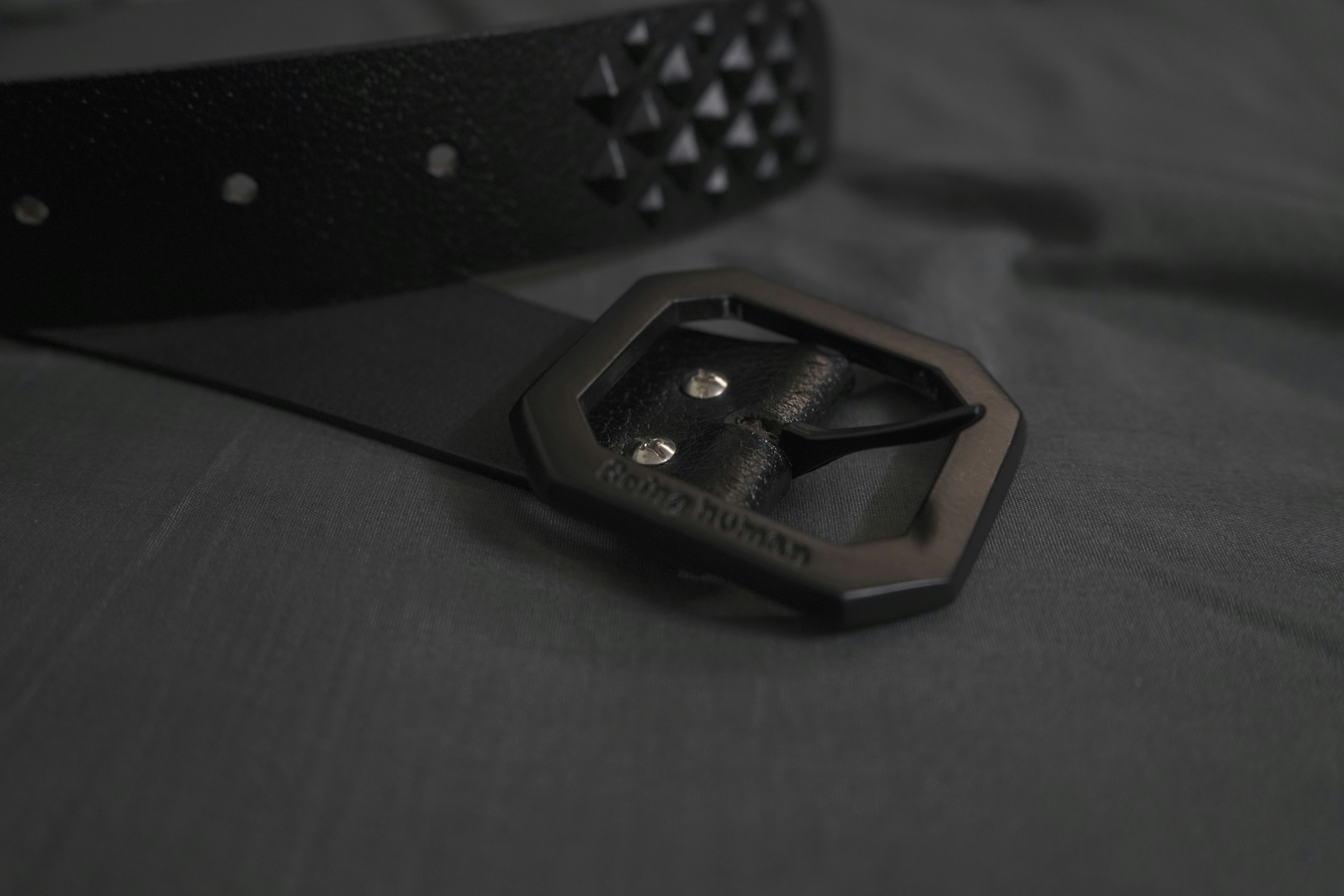 A black leather belt with a hexagonal buckle and pyramid studs on a grey surface