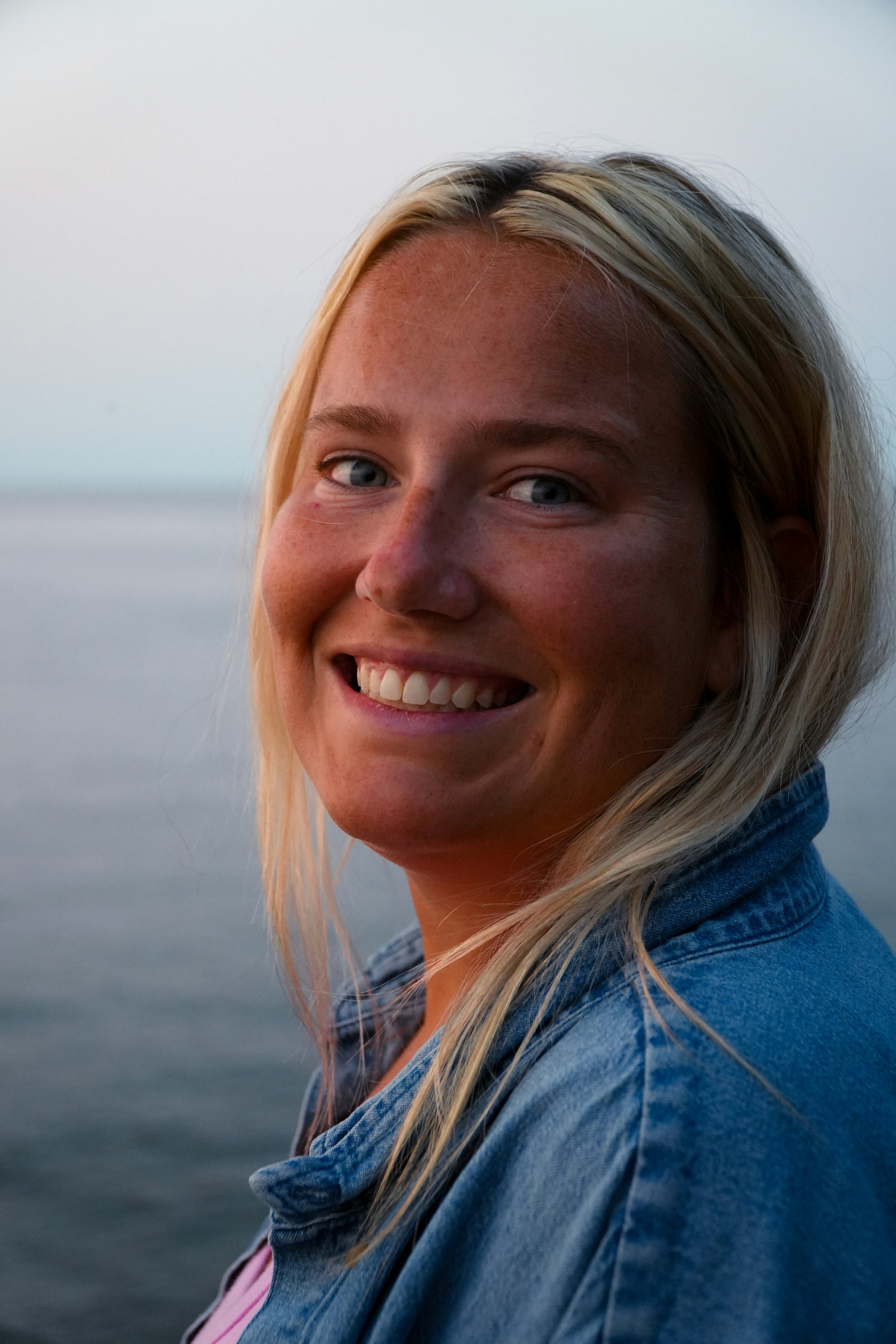 A smiling woman with blonde hair wearing a denim jacket by the water.