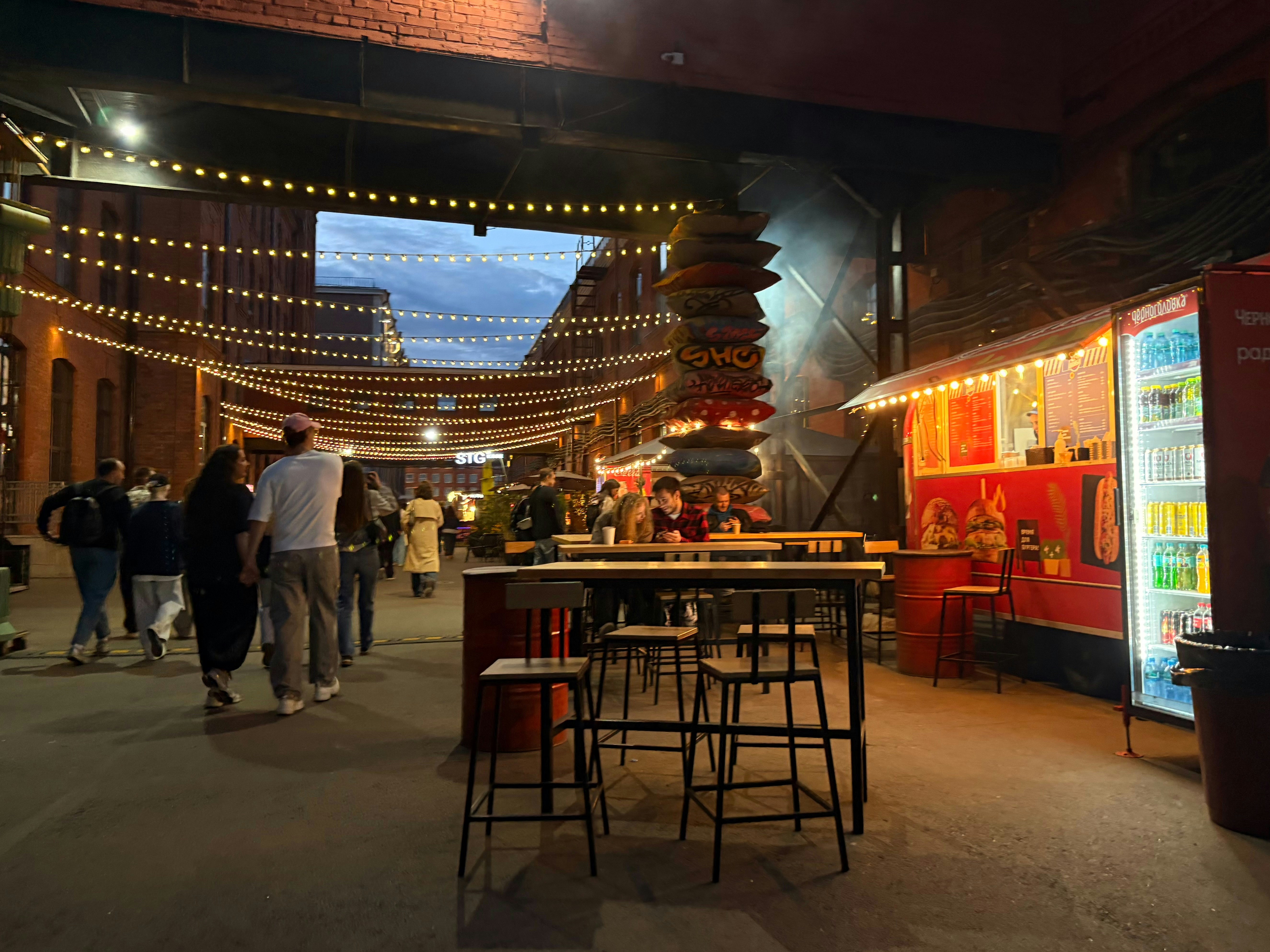 People walking and sitting at tables near a red food truck under string lights