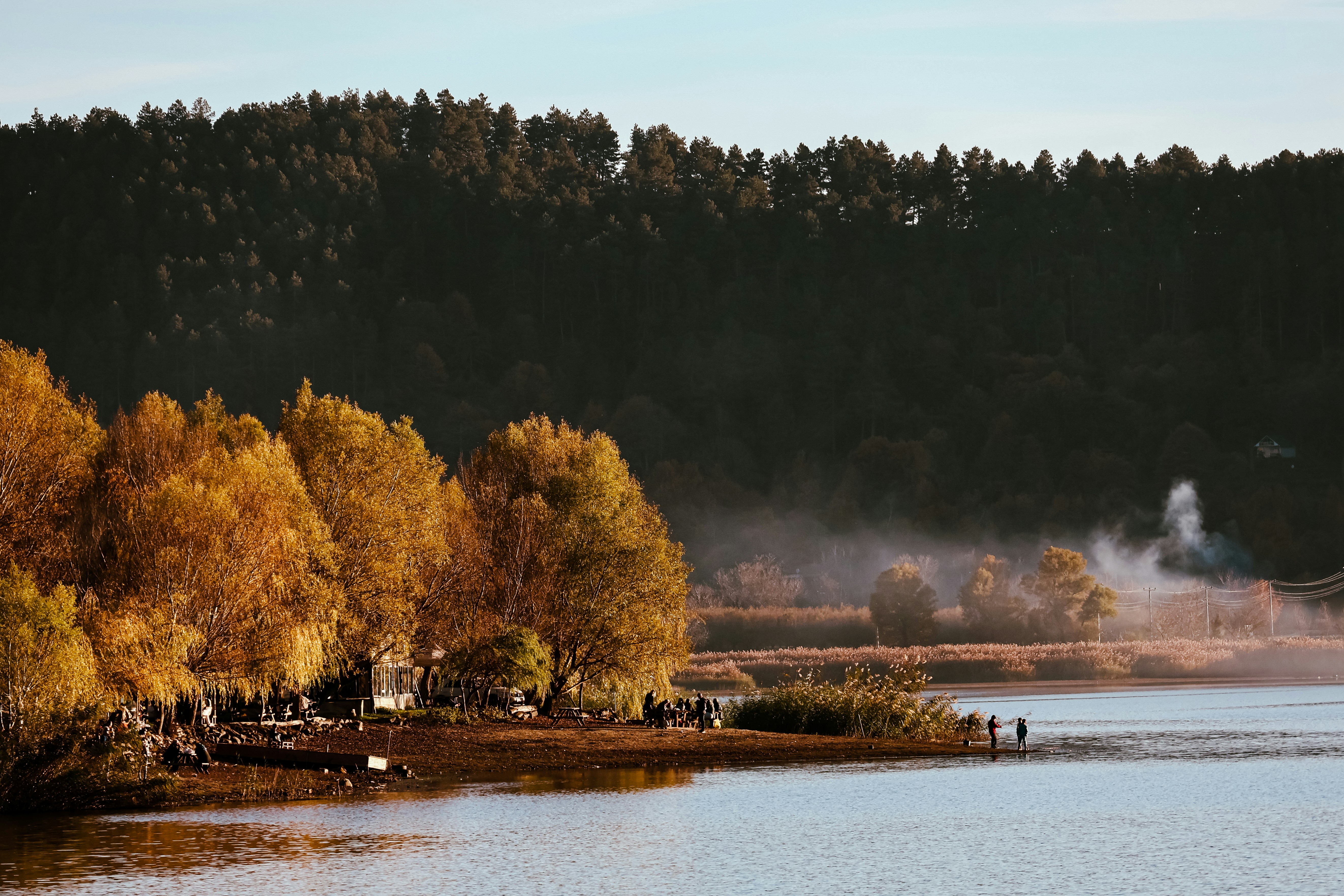People fishing on a muddy bank beside golden autumn trees and a misty lake