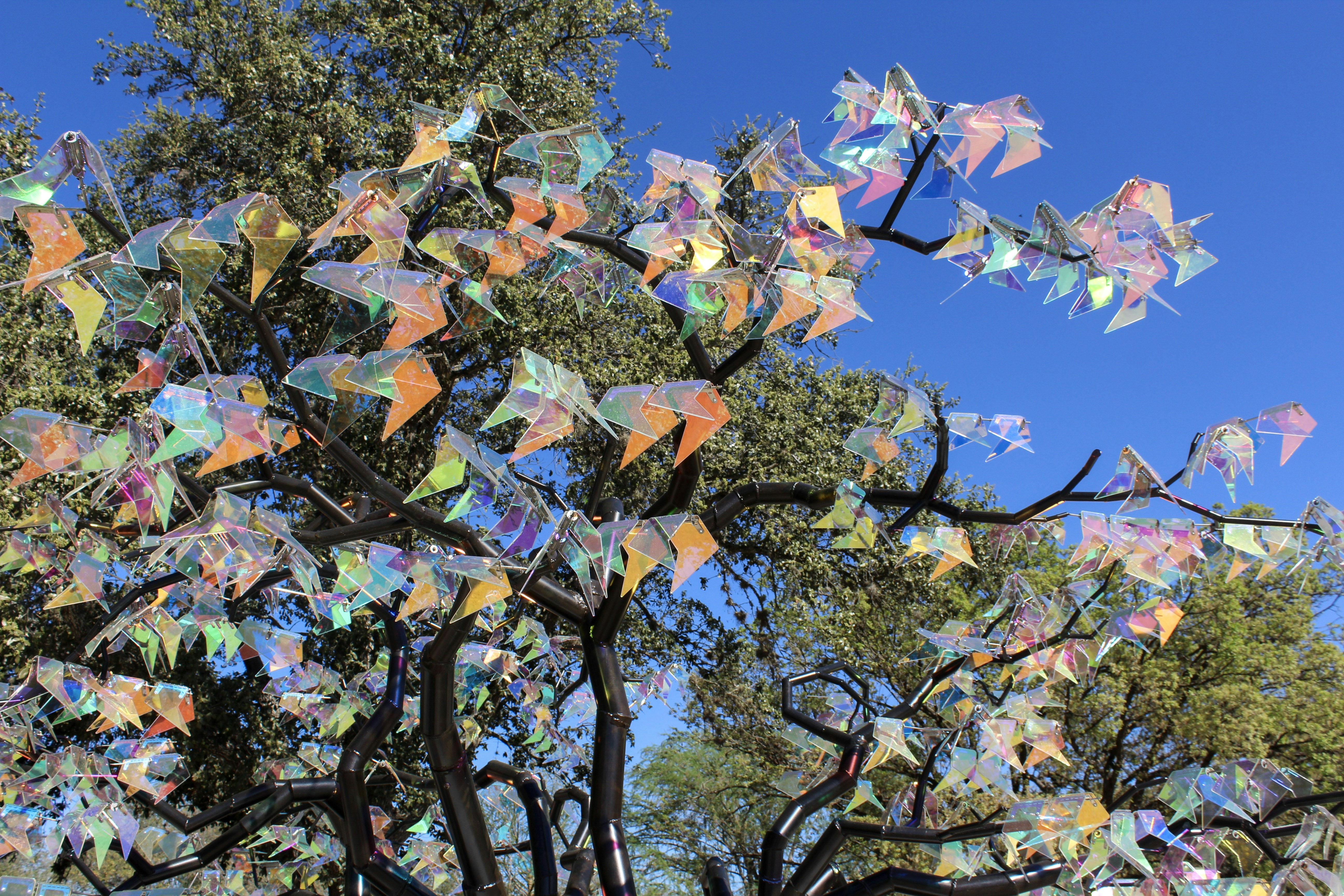 A metal tree sculpture with iridescent geometric leaves against a clear blue sky
