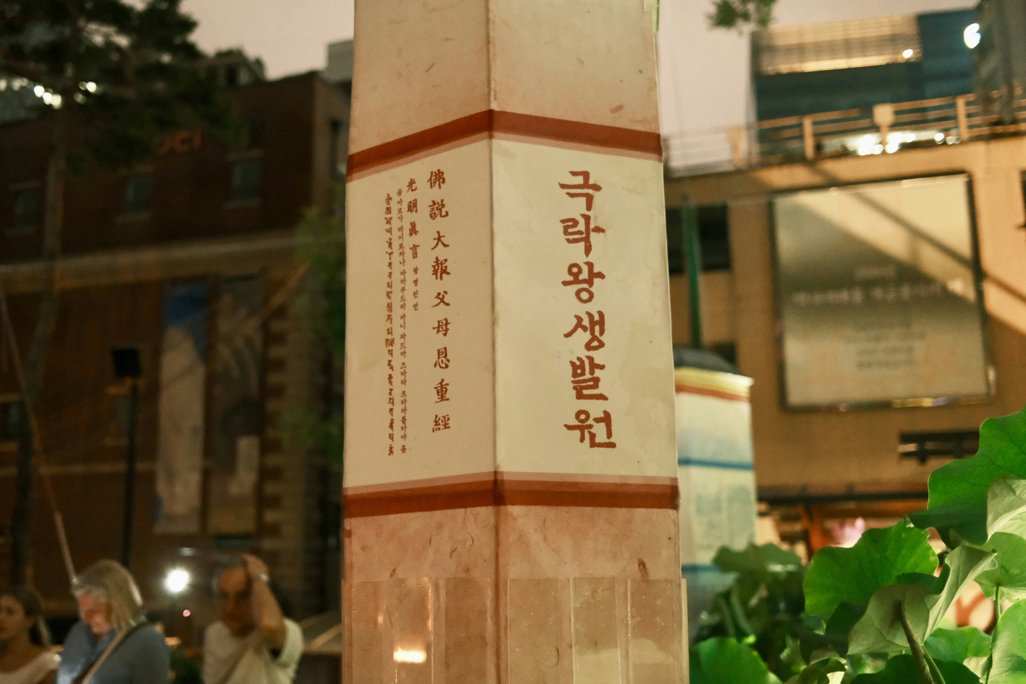 A tall illuminated pillar with Korean text in a city square at night
