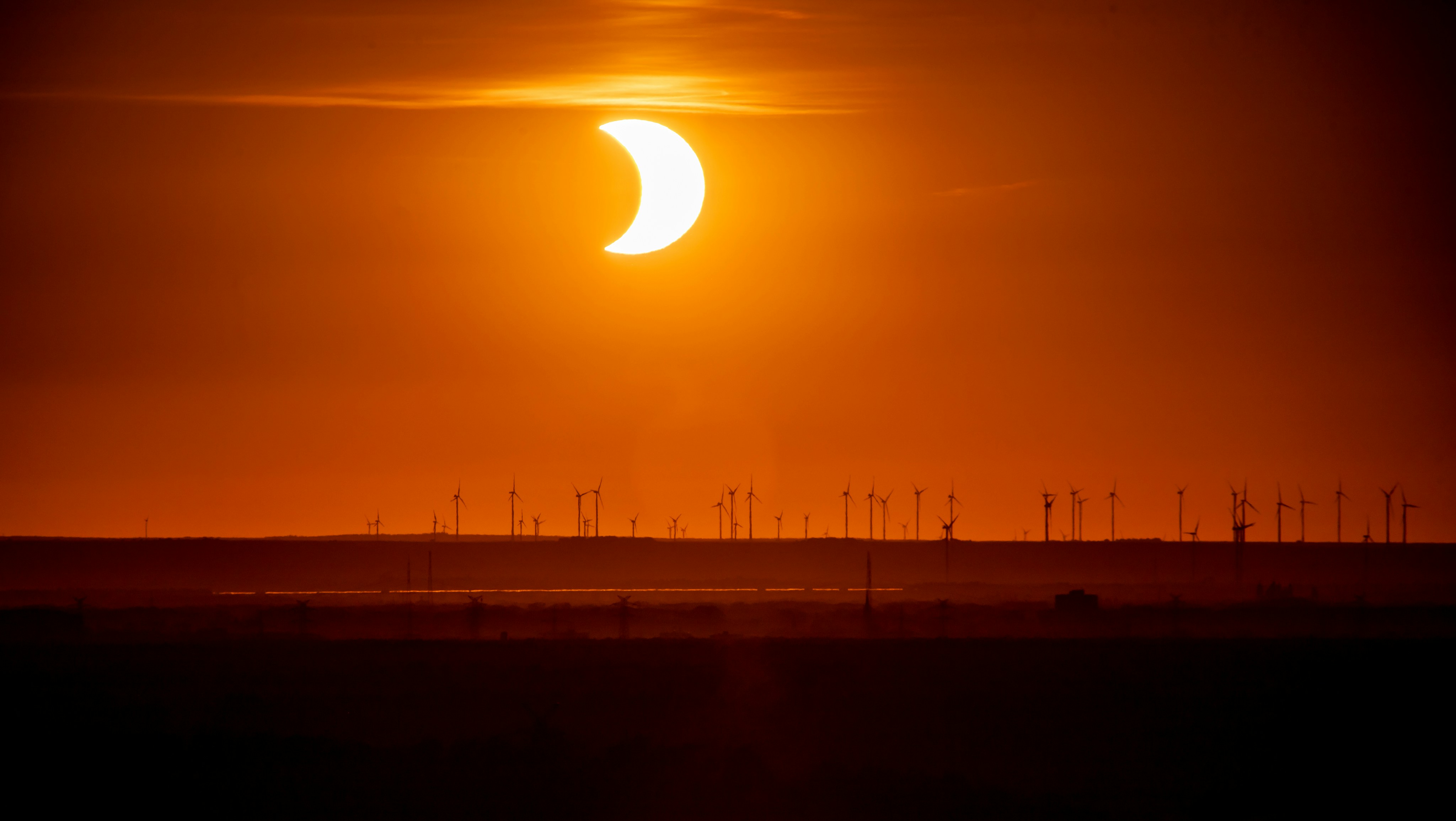 A partial solar eclipse above a line of wind turbines in a warm orange sky.