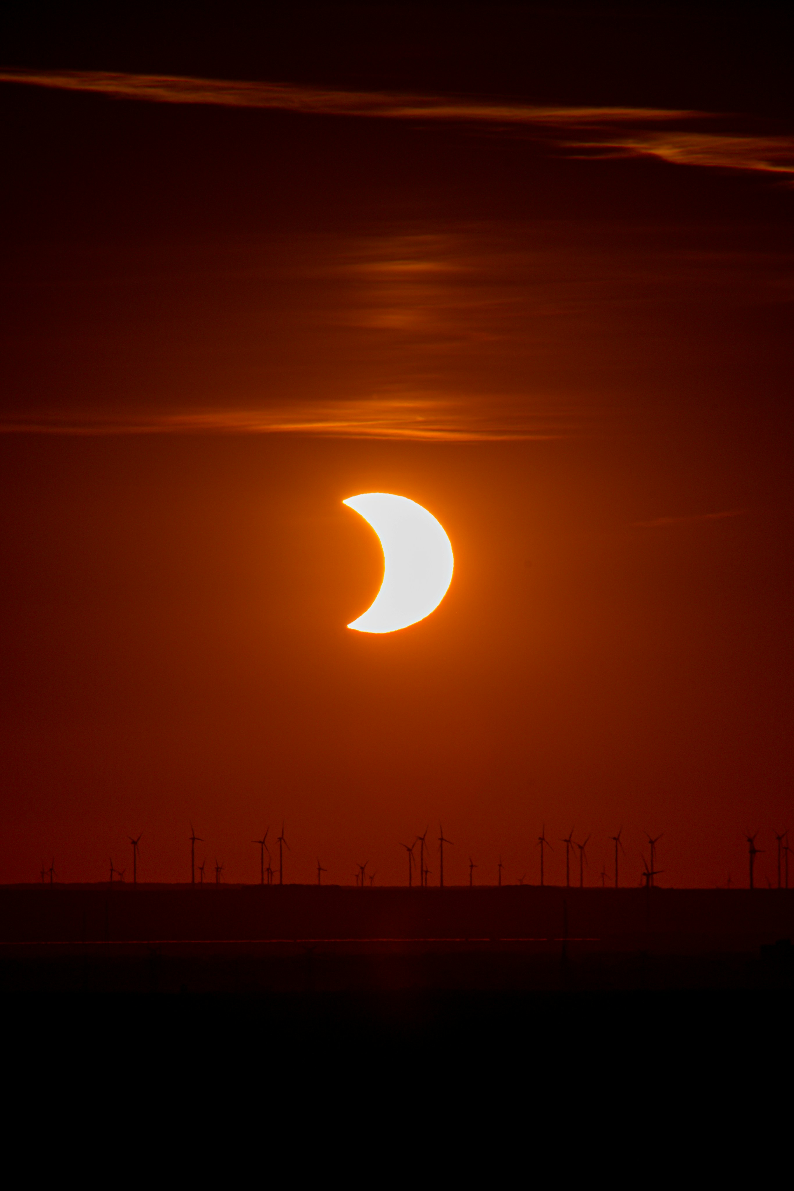 A partial solar eclipse above a silhouette of wind turbines at low light.