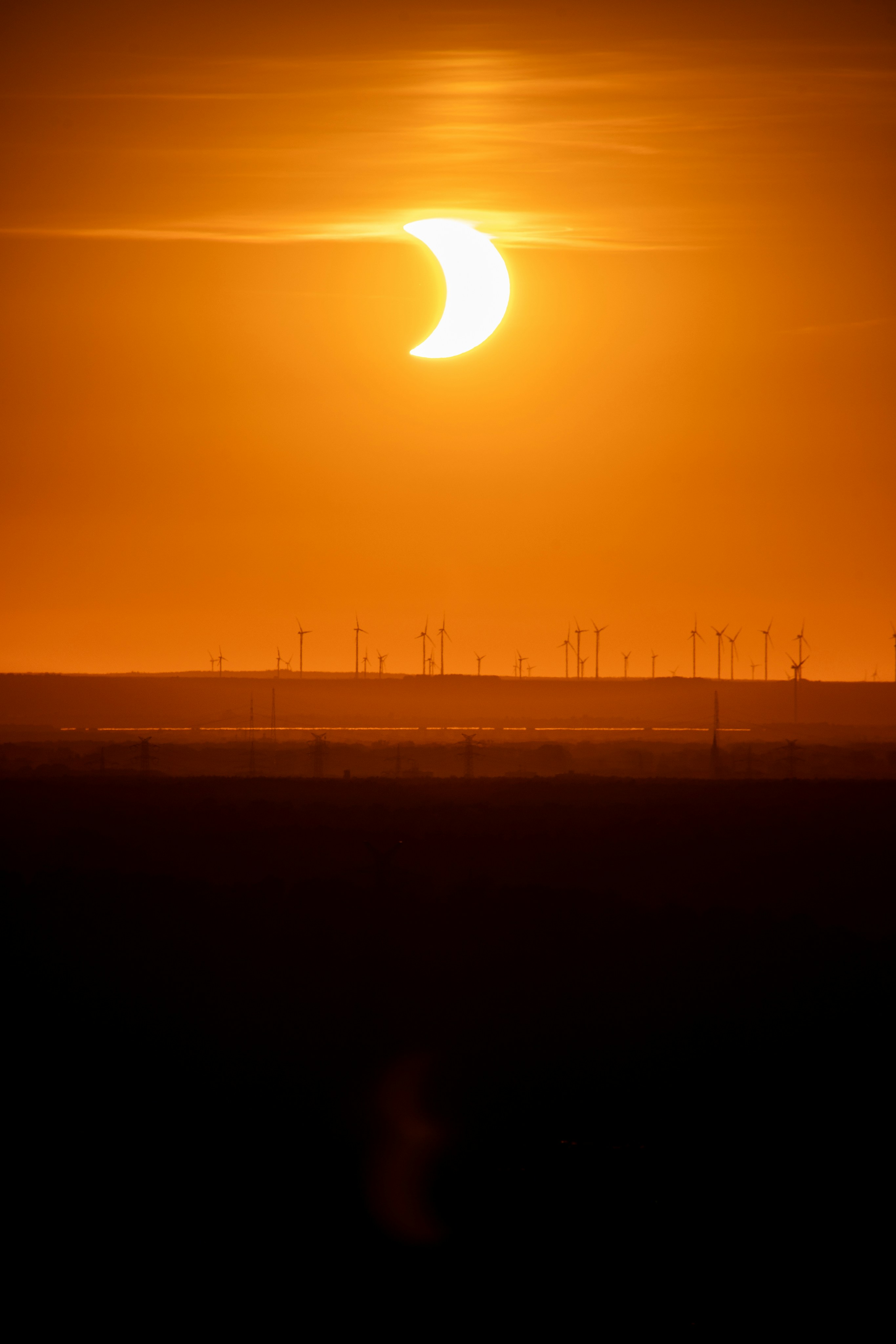 A partial solar eclipse above a field of wind turbines in a warm orange sky.