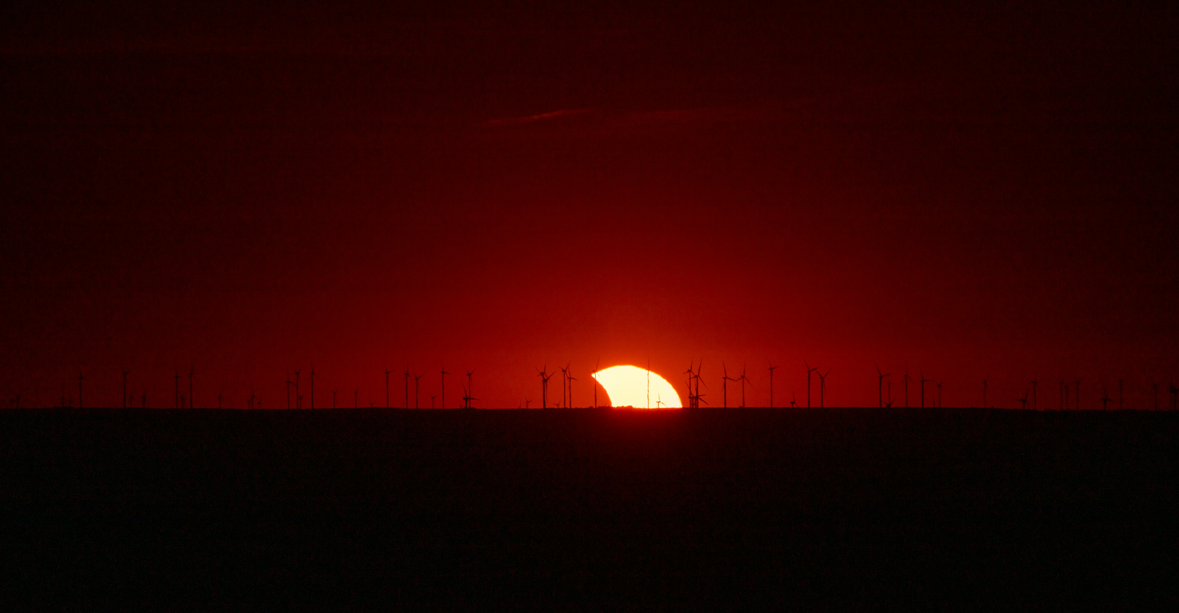 A partial solar eclipse behind a line of wind turbines at low light.