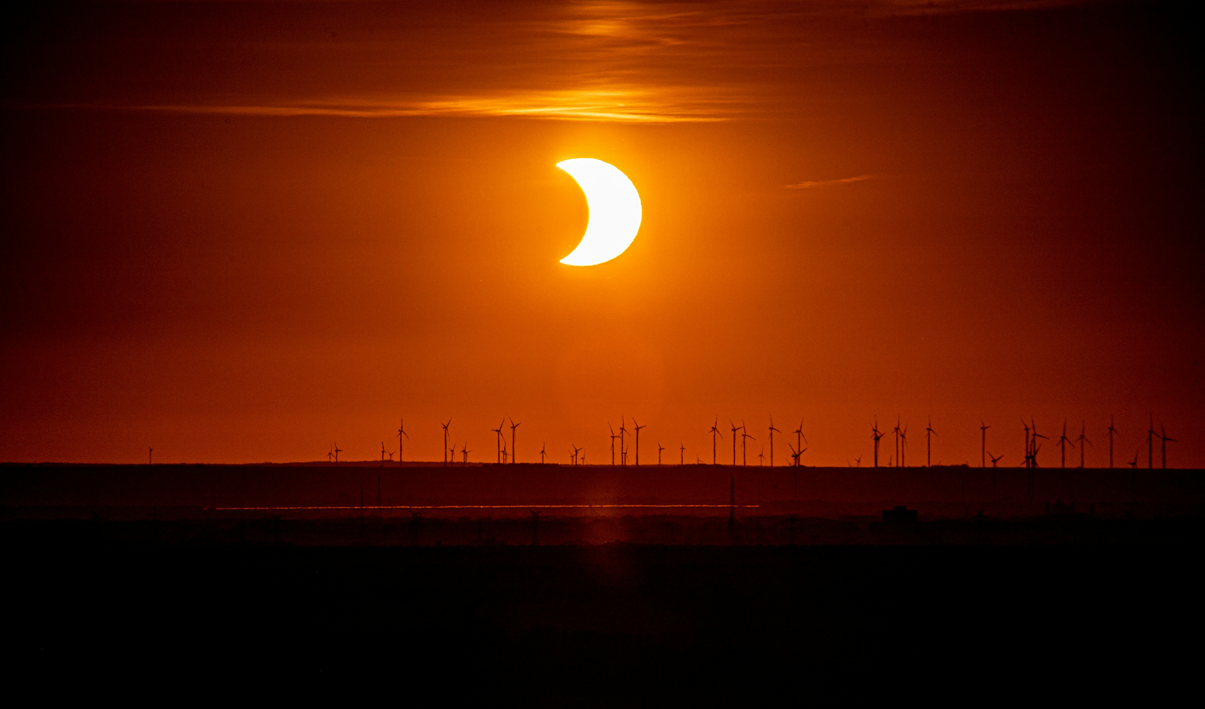A partial solar eclipse above a line of wind turbines at low sun