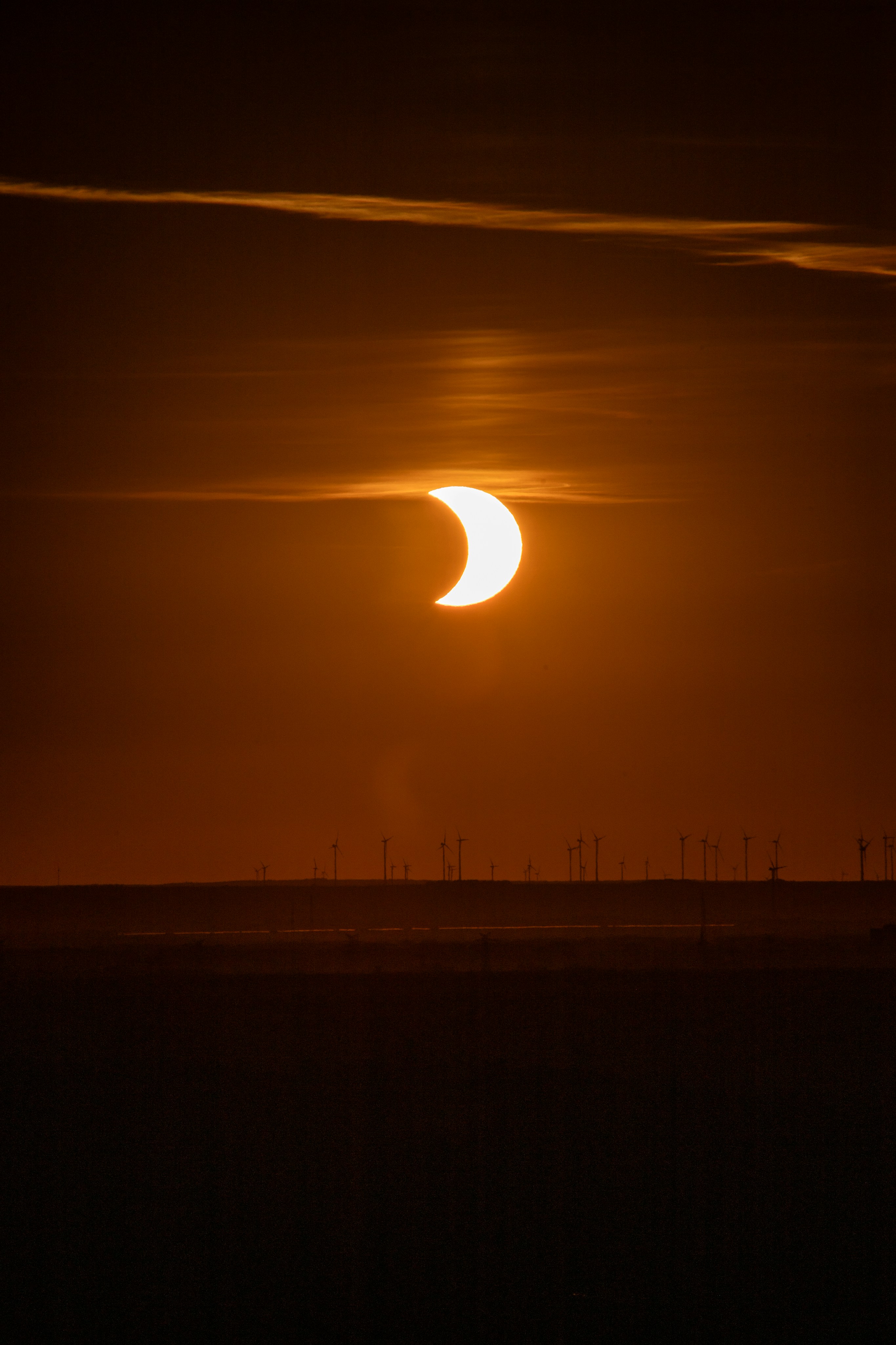 A partial solar eclipse above a dark horizon with silhouettes of wind turbines.