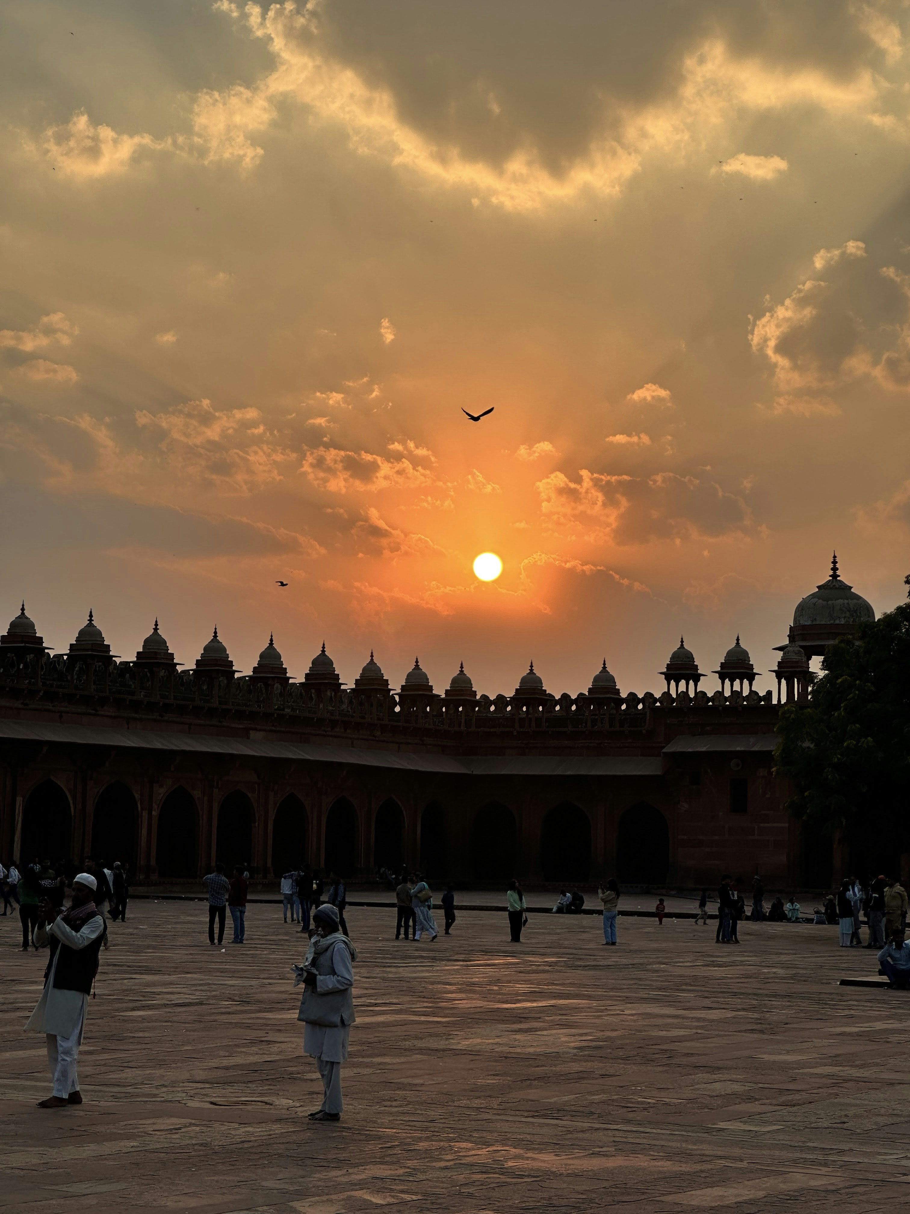 People in a courtyard before the red sandstone architecture of Fatehpur Sikri at low sun.