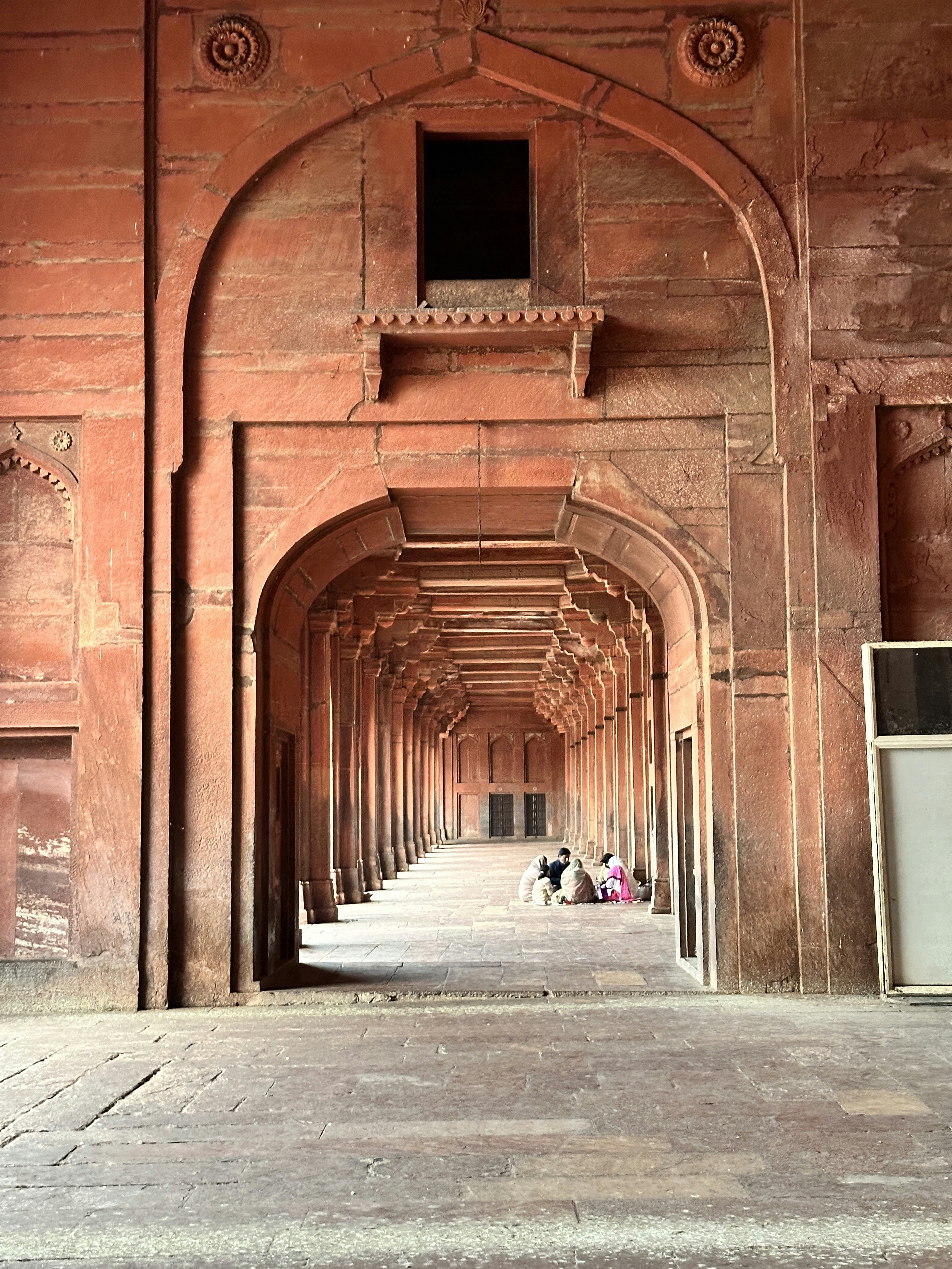 A group of people sitting on the floor within a red sandstone arched corridor.