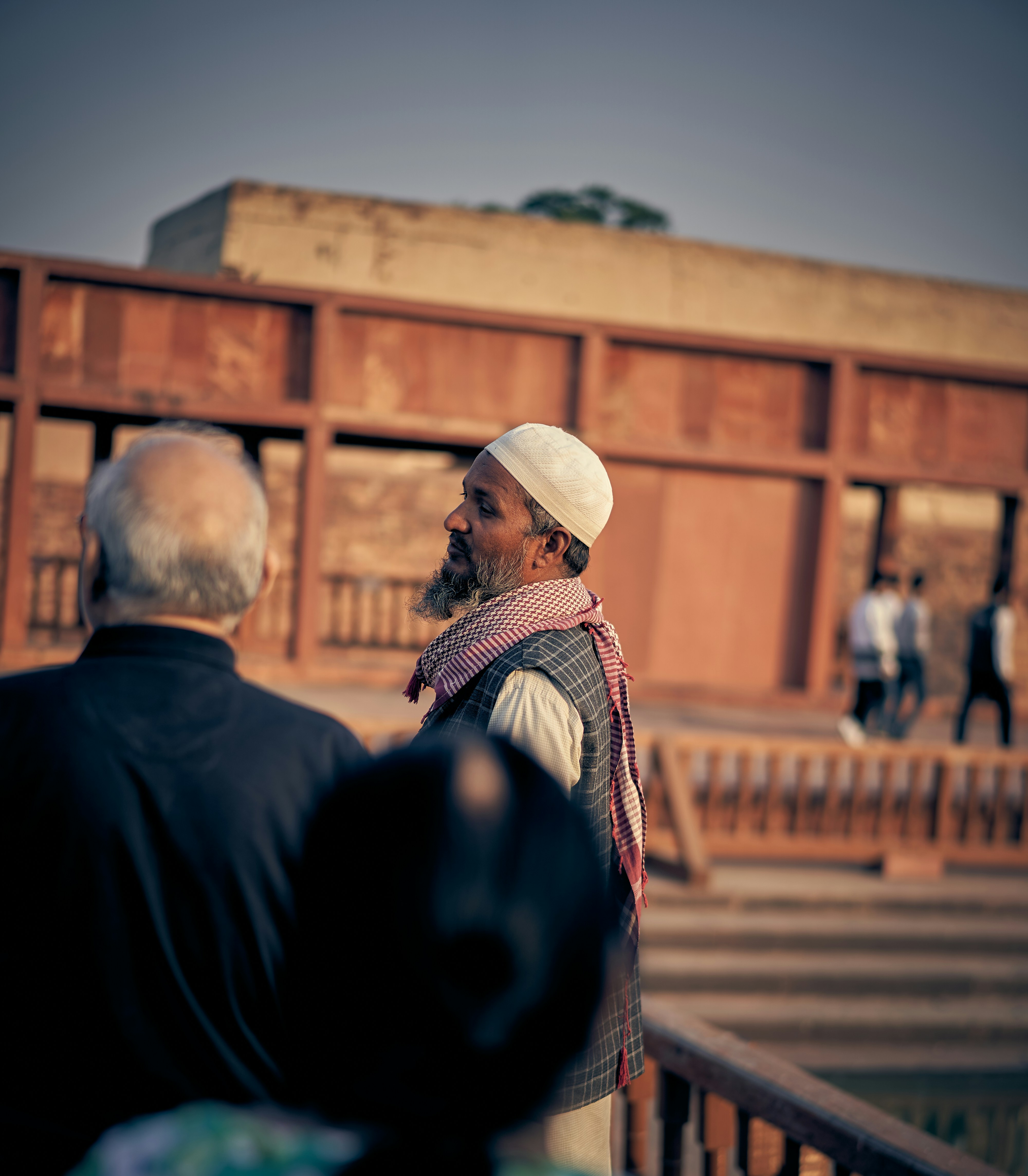 A man in a white skullcap and patterned scarf standing near a red building.
