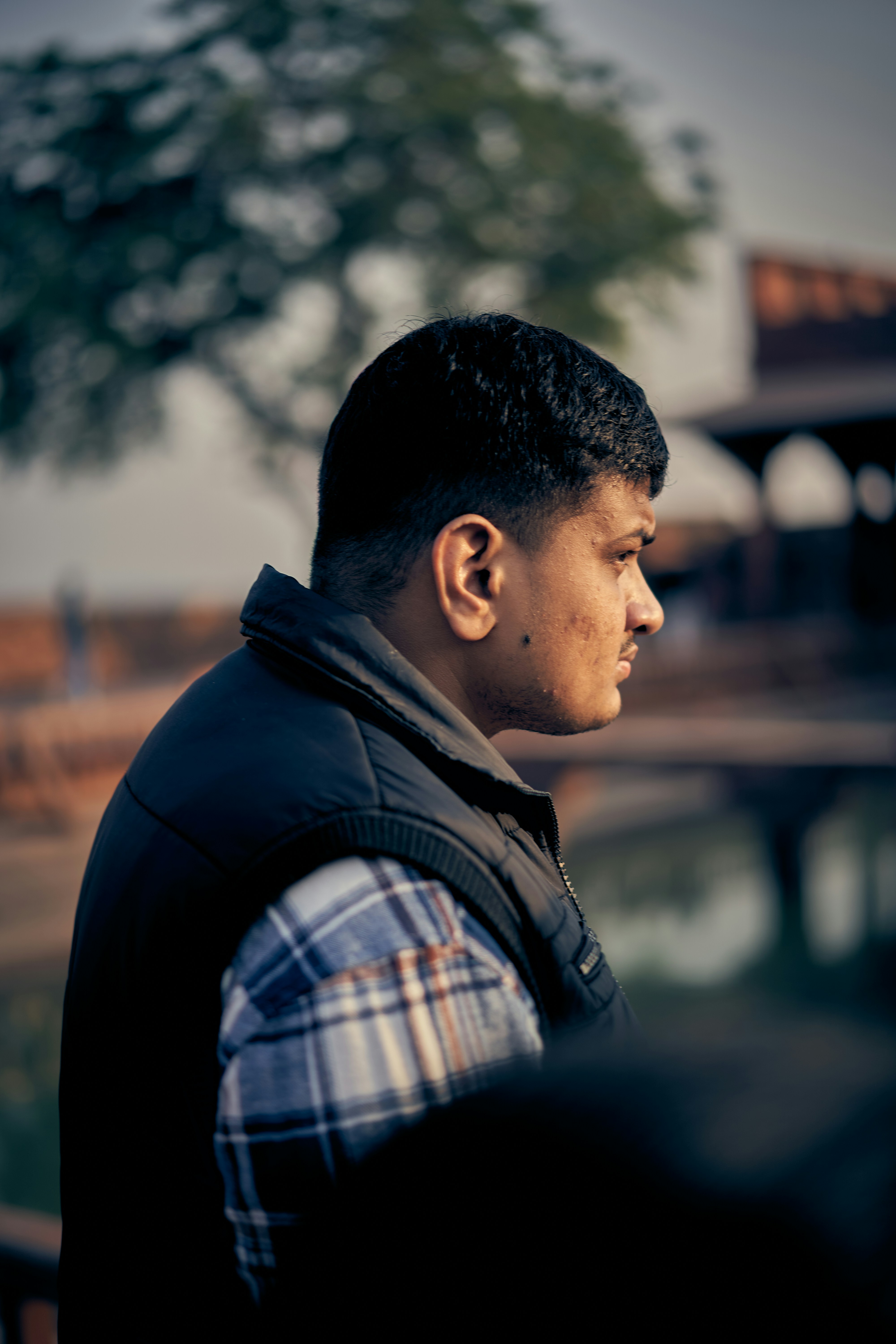 A man in a plaid shirt and black vest looking away outdoors.
