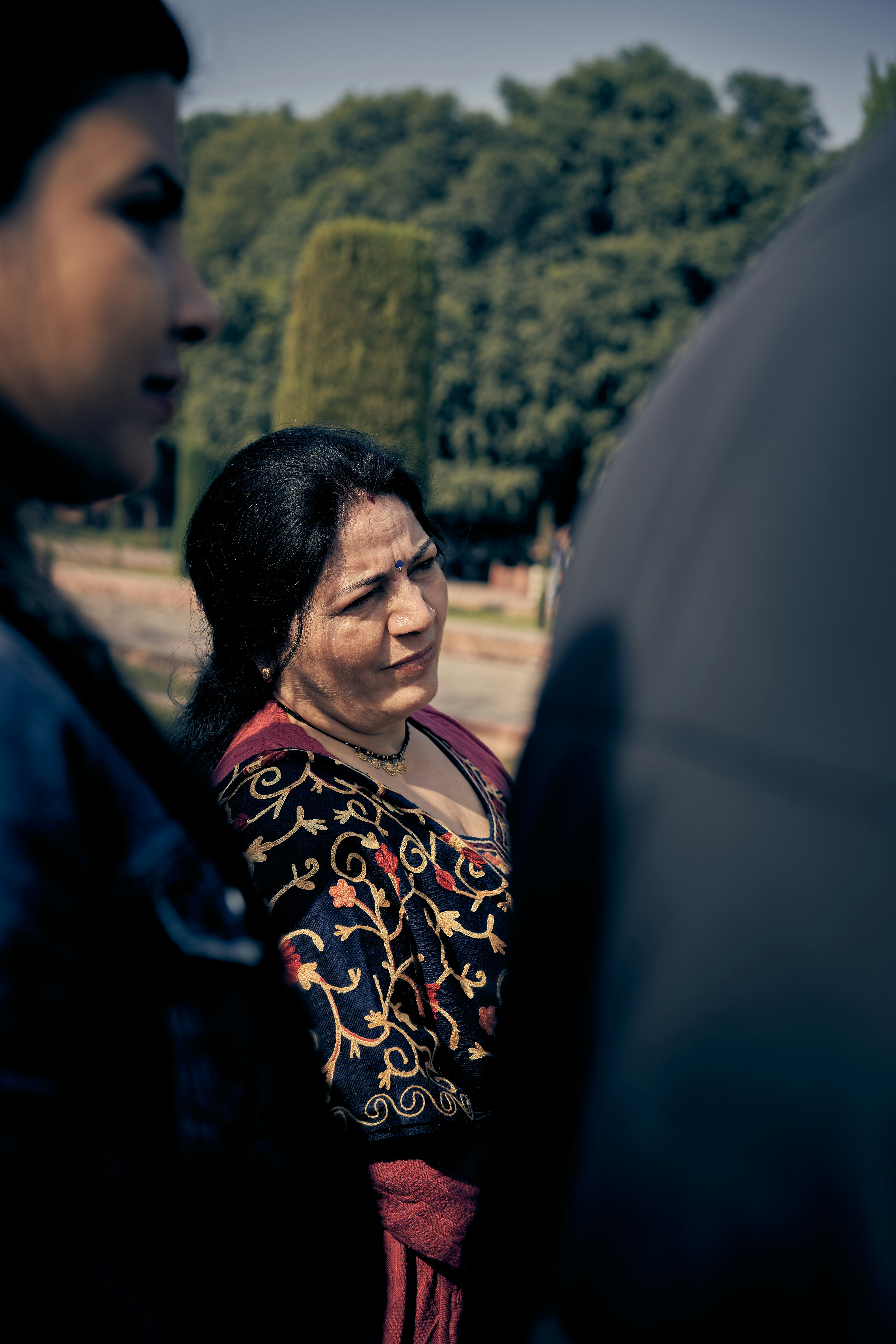 A woman in a dark embroidered shawl standing outdoors near trees.