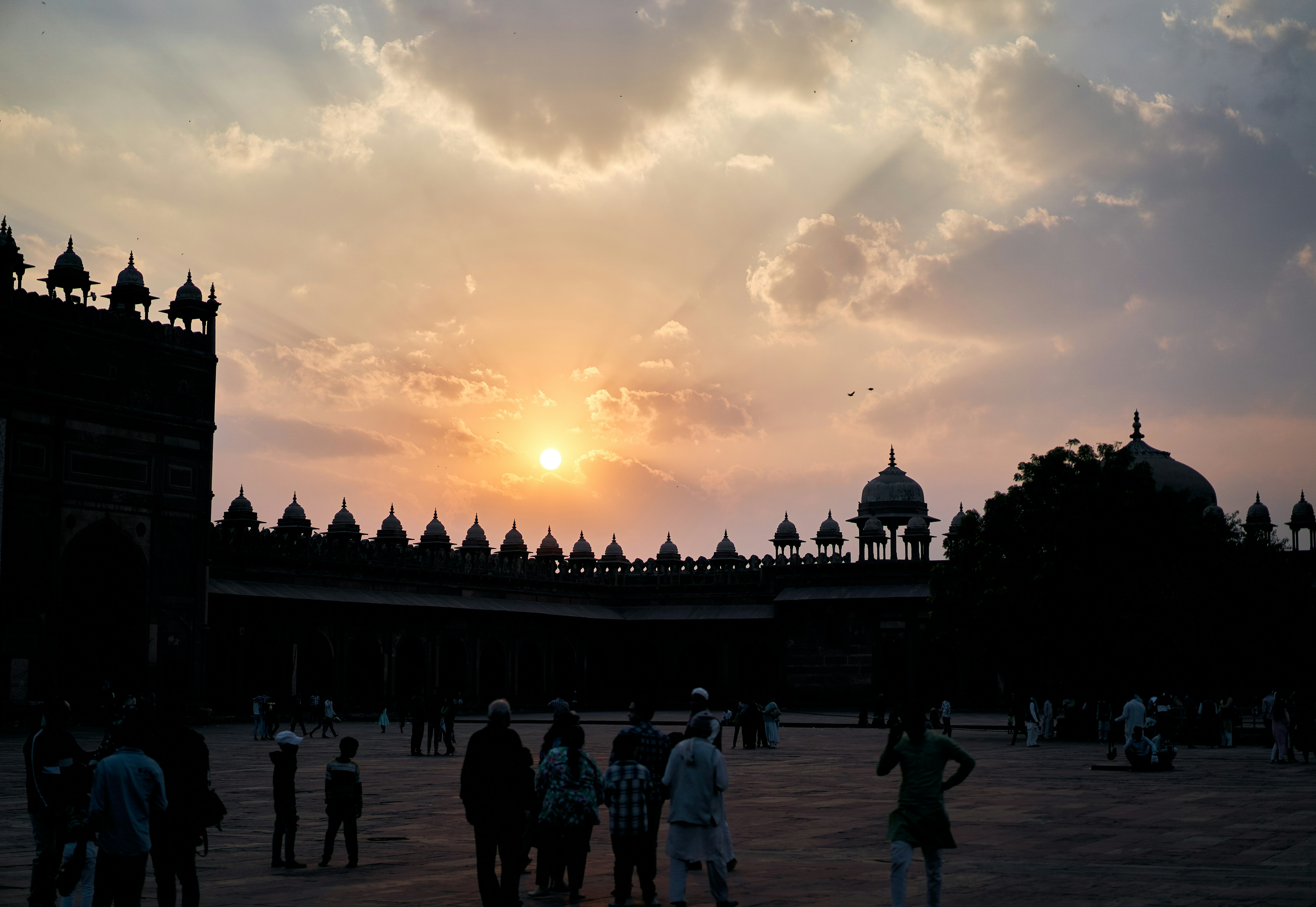 Silhouettes of people in a courtyard before a domed building under a golden sky.