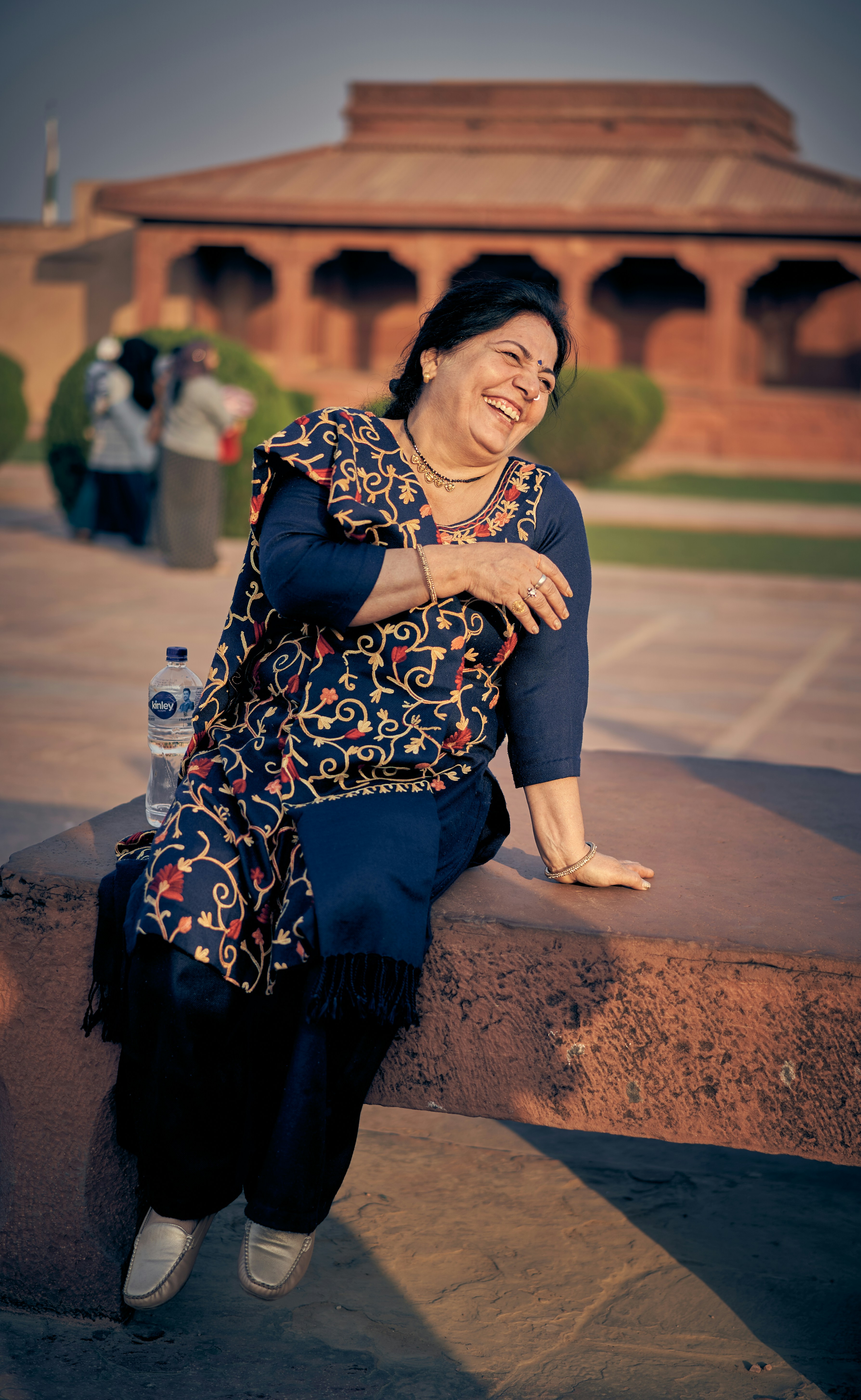 A laughing woman in a dark patterned tunic sitting on a stone ledge.