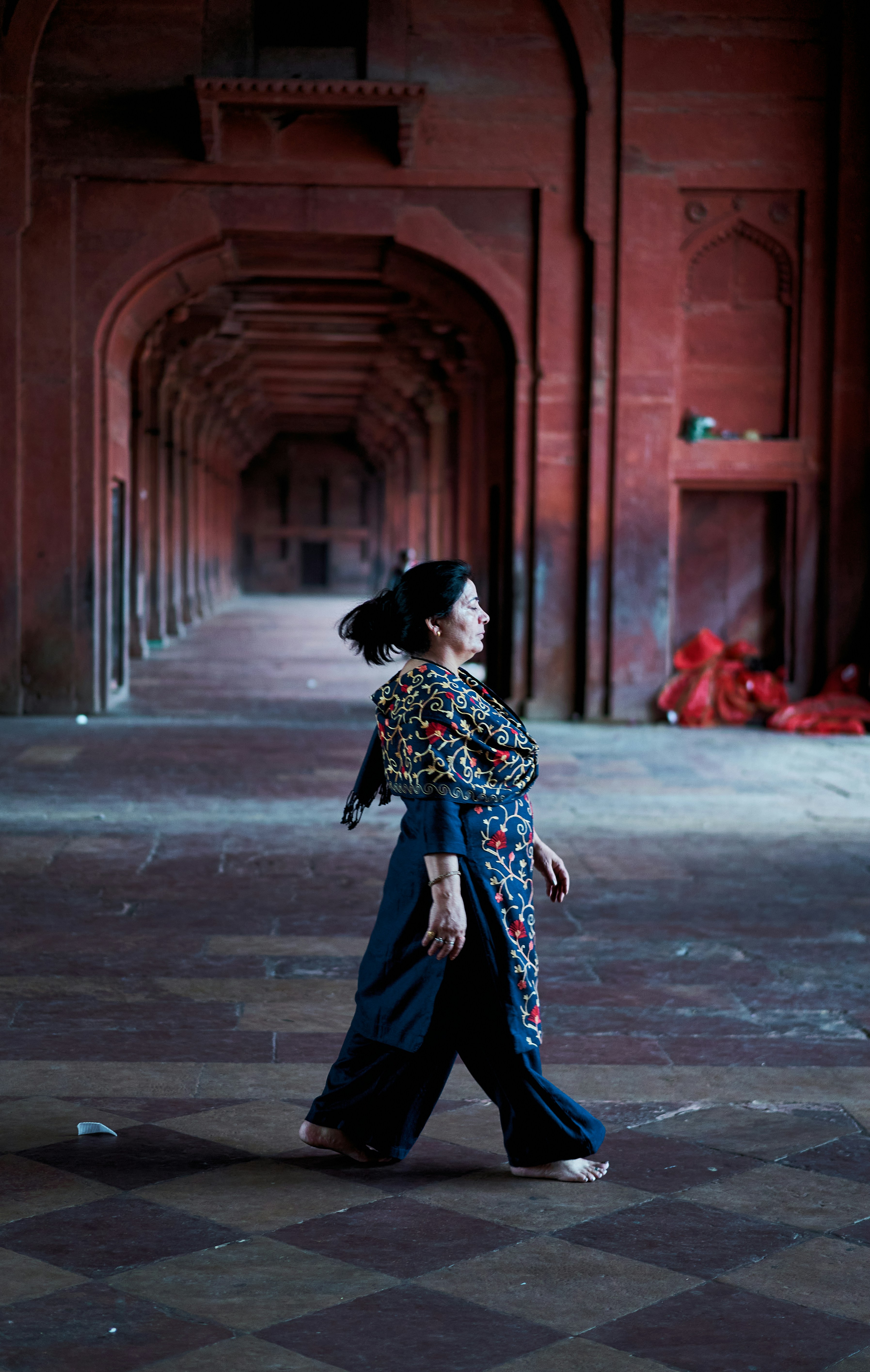 A woman in a dark blue embroidered tunic walking barefoot through a red stone corridor.