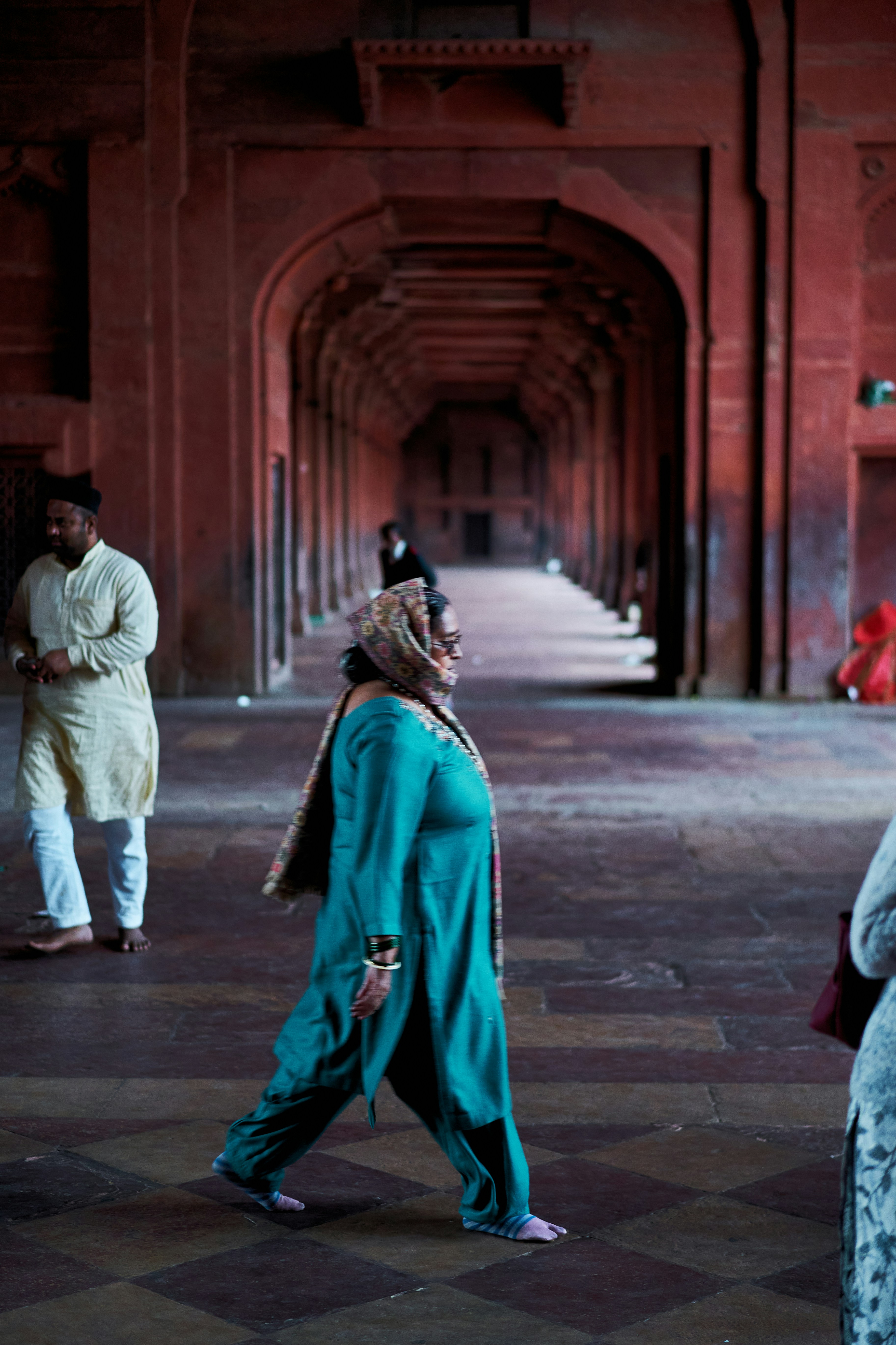 A woman in a teal tunic walking barefoot through a red sandstone corridor.