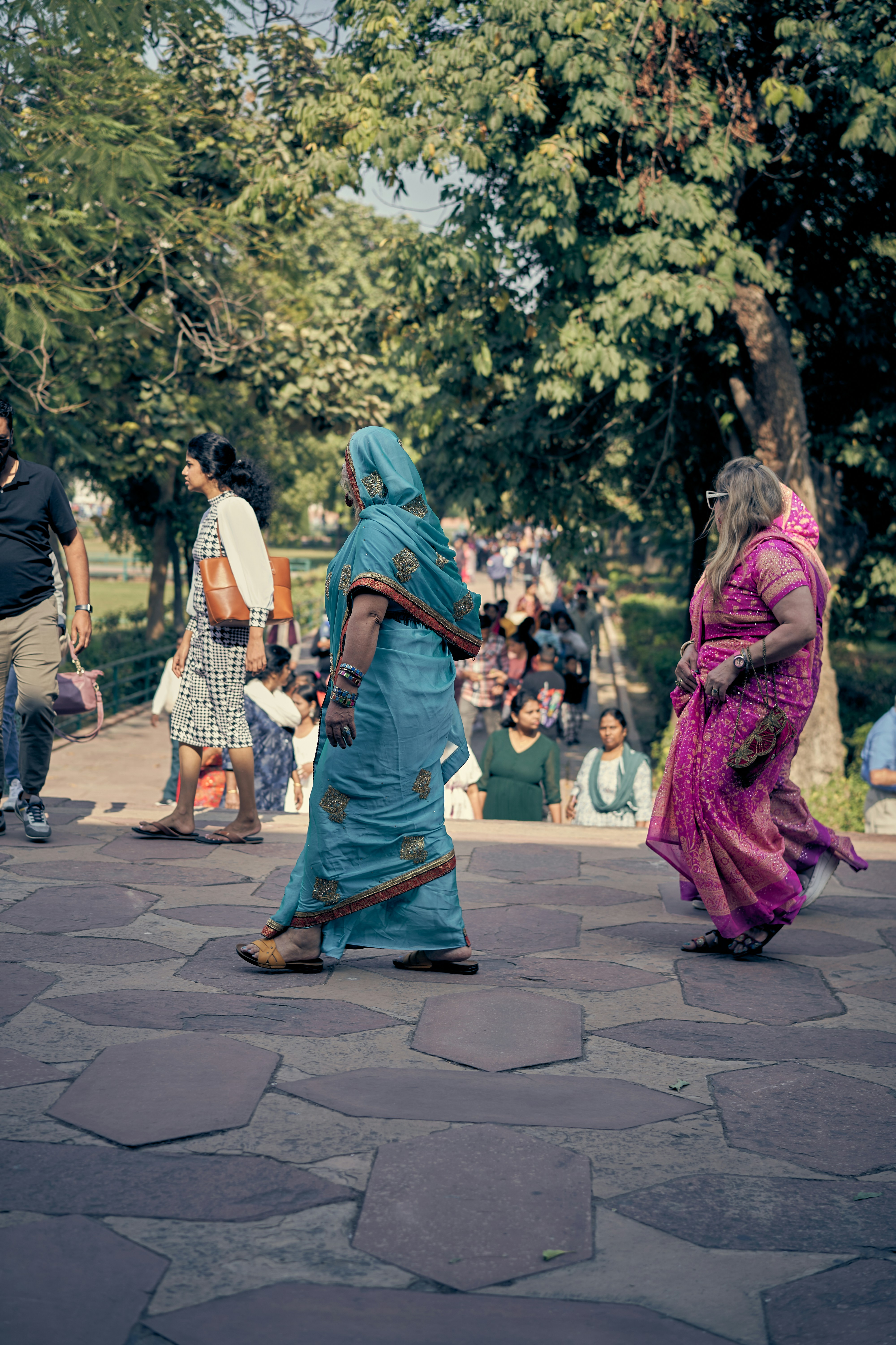 People walking on a stone path in a park shaded by large trees.