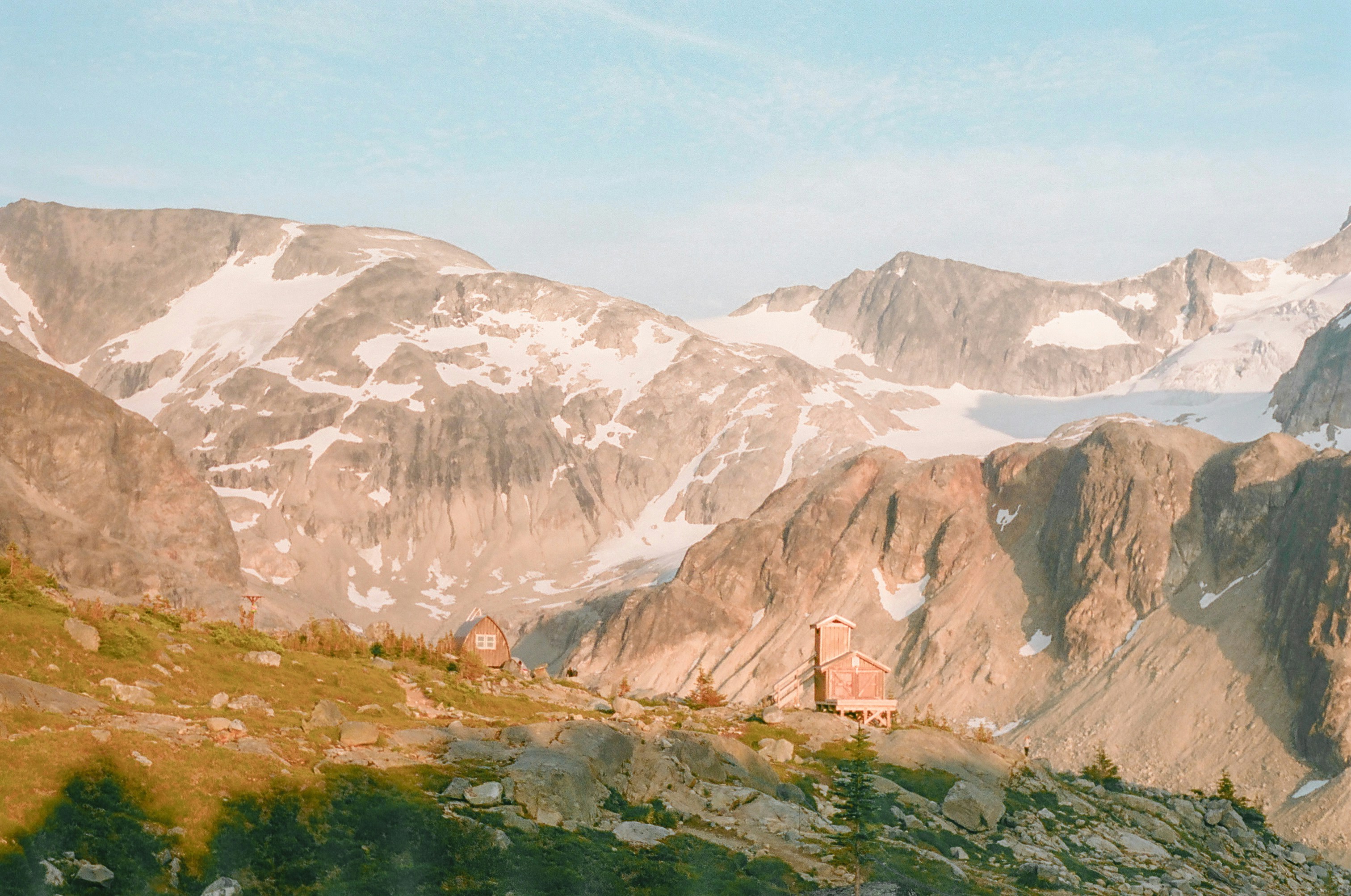 Two small wooden cabins on a rocky mountain slope under a clear sky.