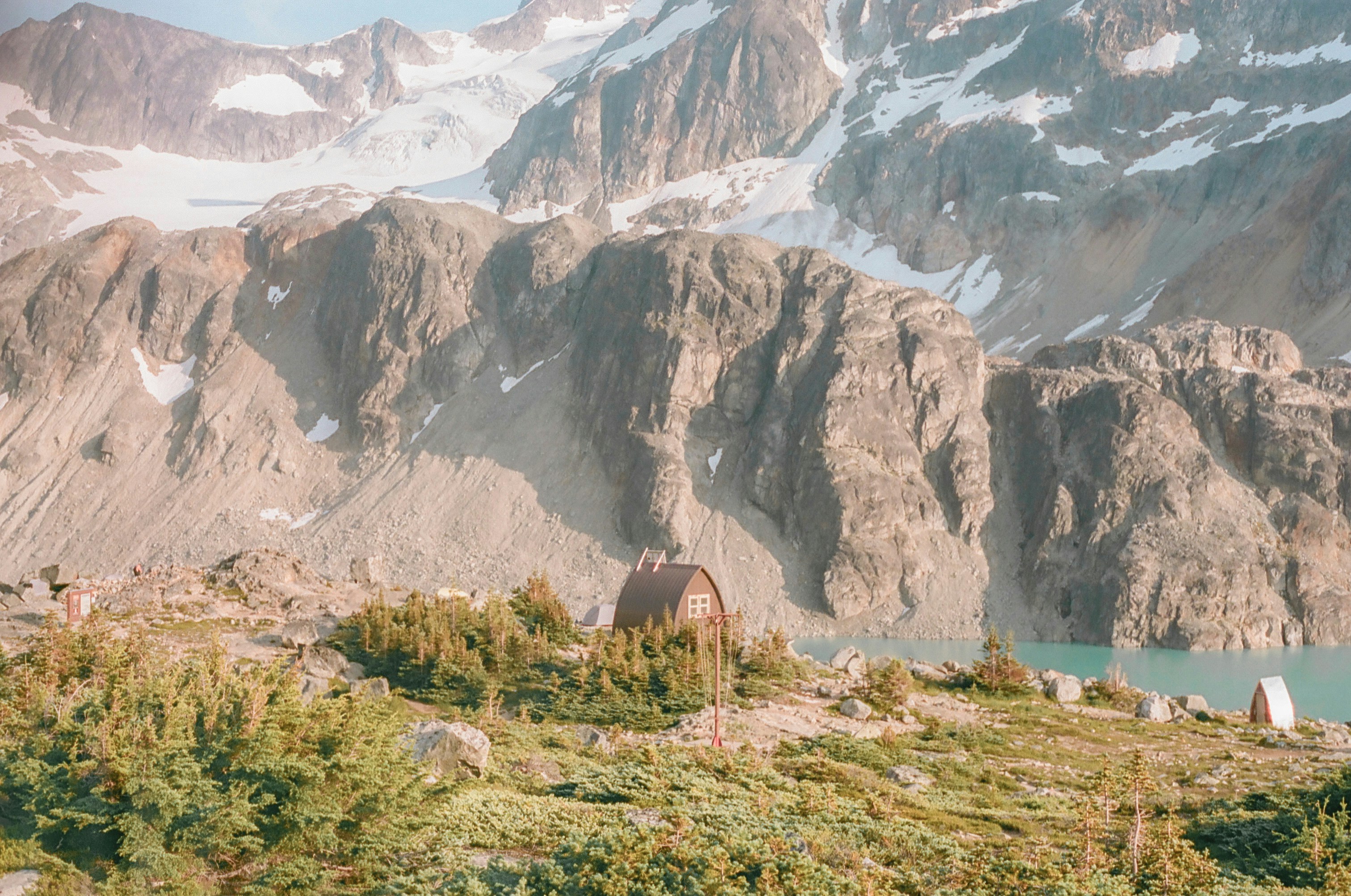 A small cabin near a turquoise alpine lake beneath rugged, snow-dusted mountain peaks.