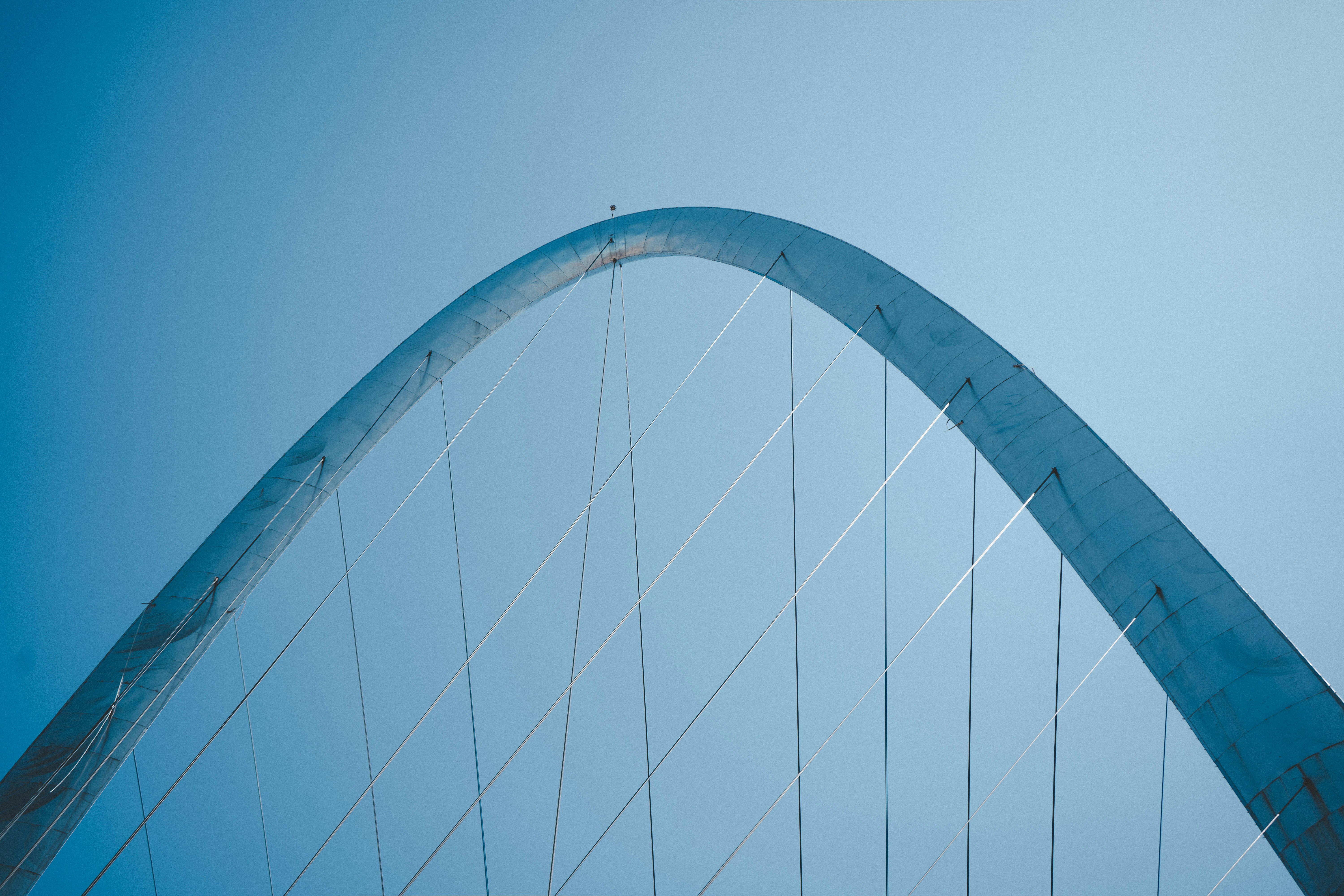 A curved metal bridge arch with suspension cables against a clear blue sky.