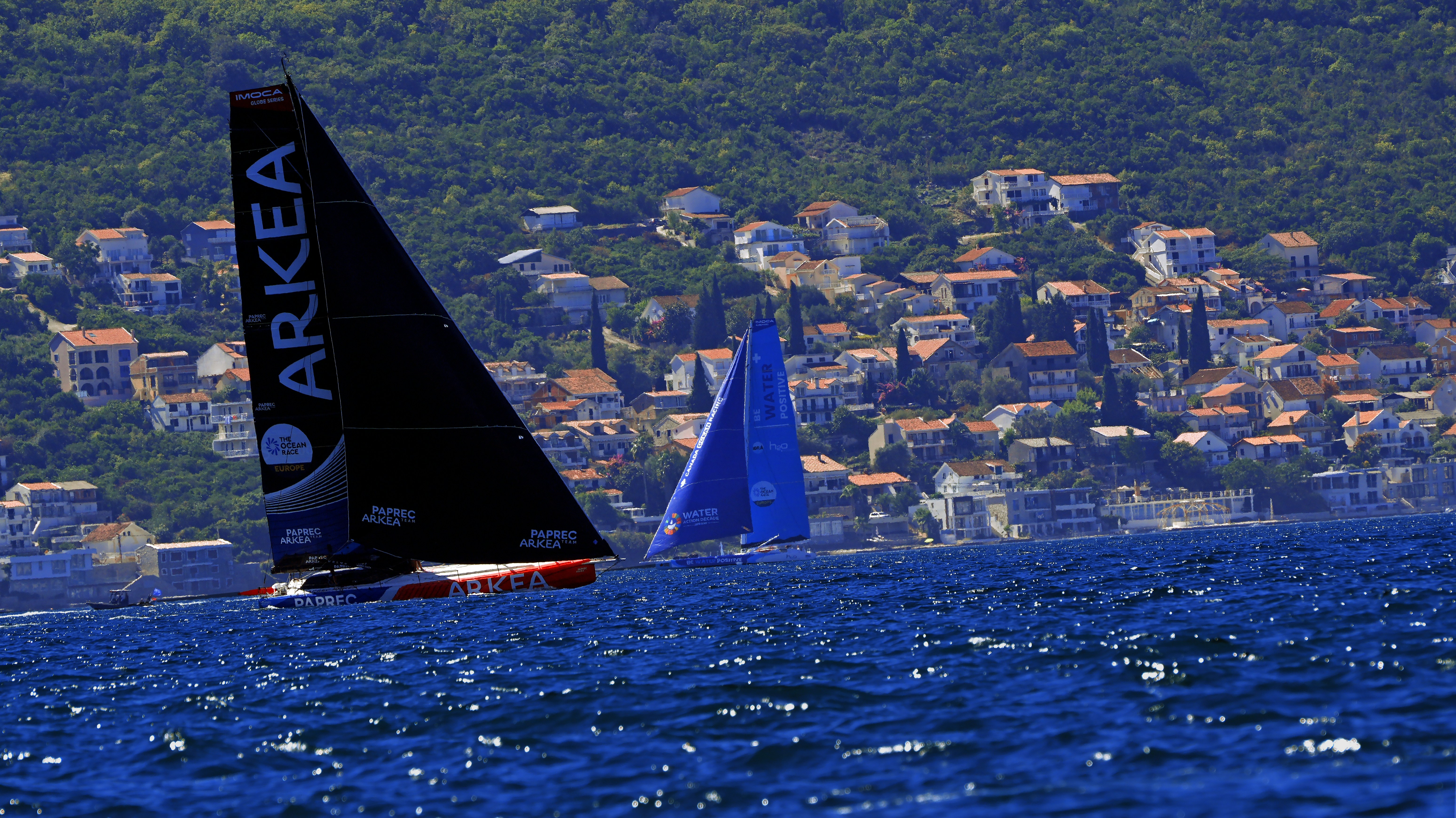 Two racing sailboats on blue water near a hillside town with ma…