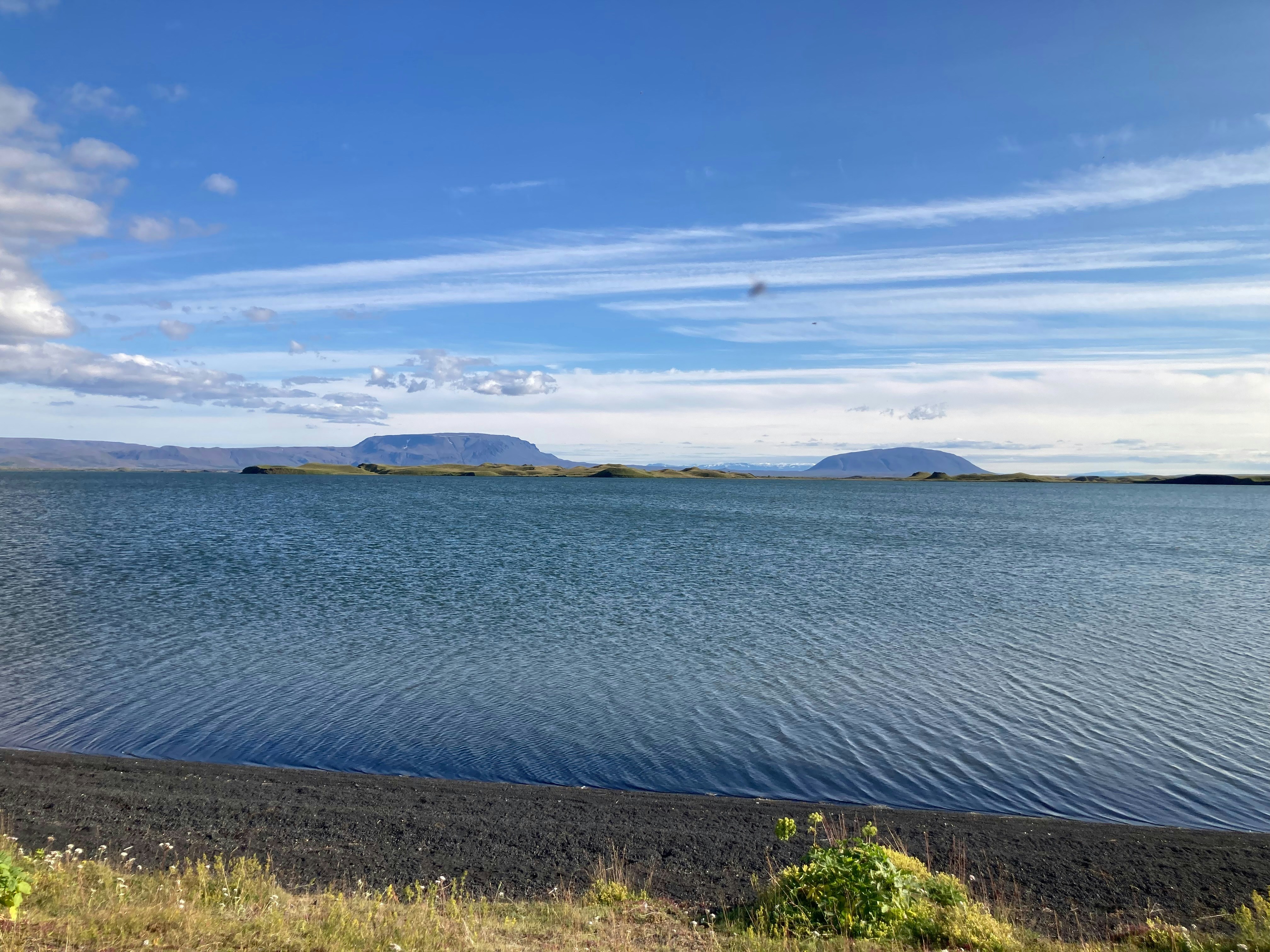 A dark volcanic sand beach bordering a wide lake with distant mountains