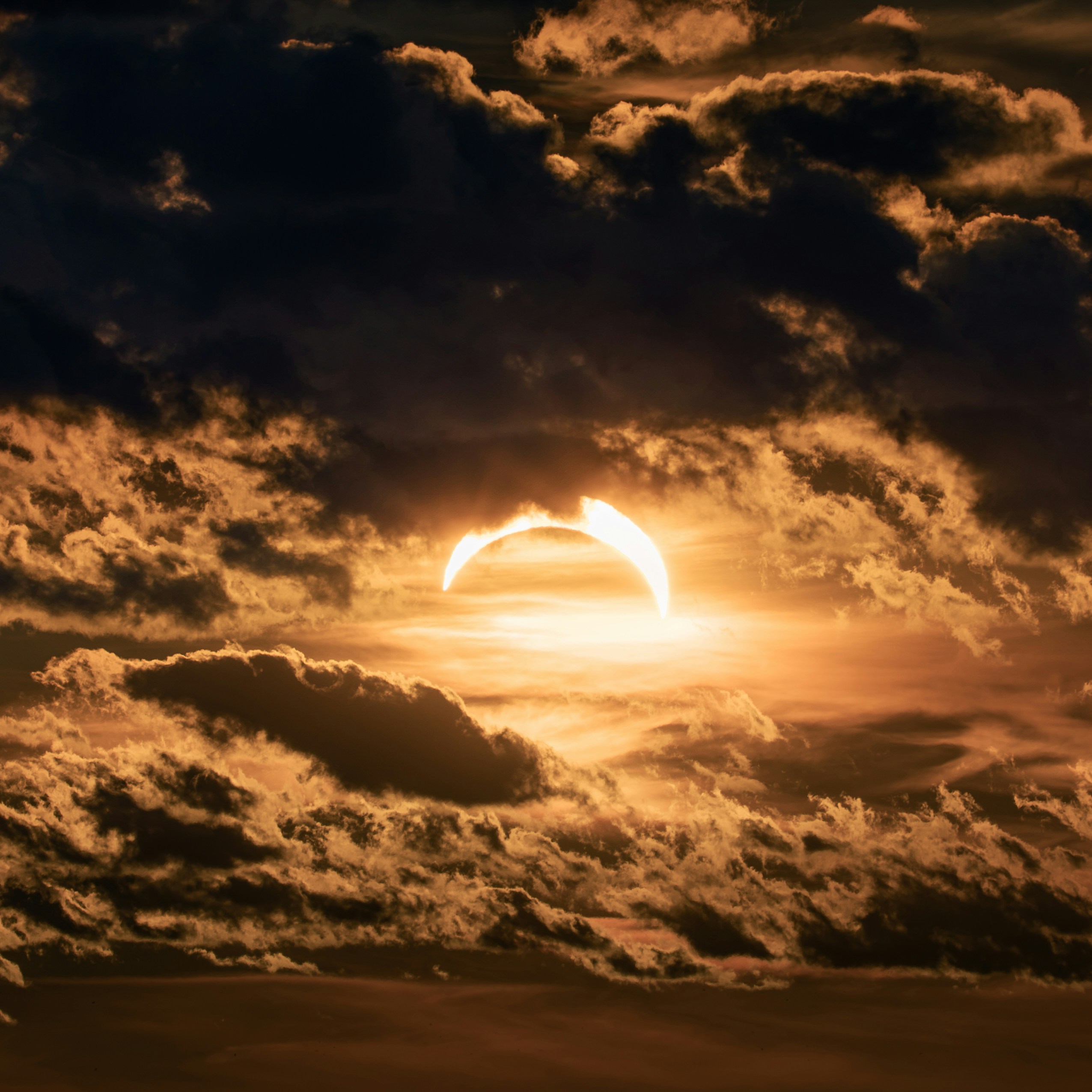 A partial solar eclipse glowing behind dark, textured clouds in a warm sky.