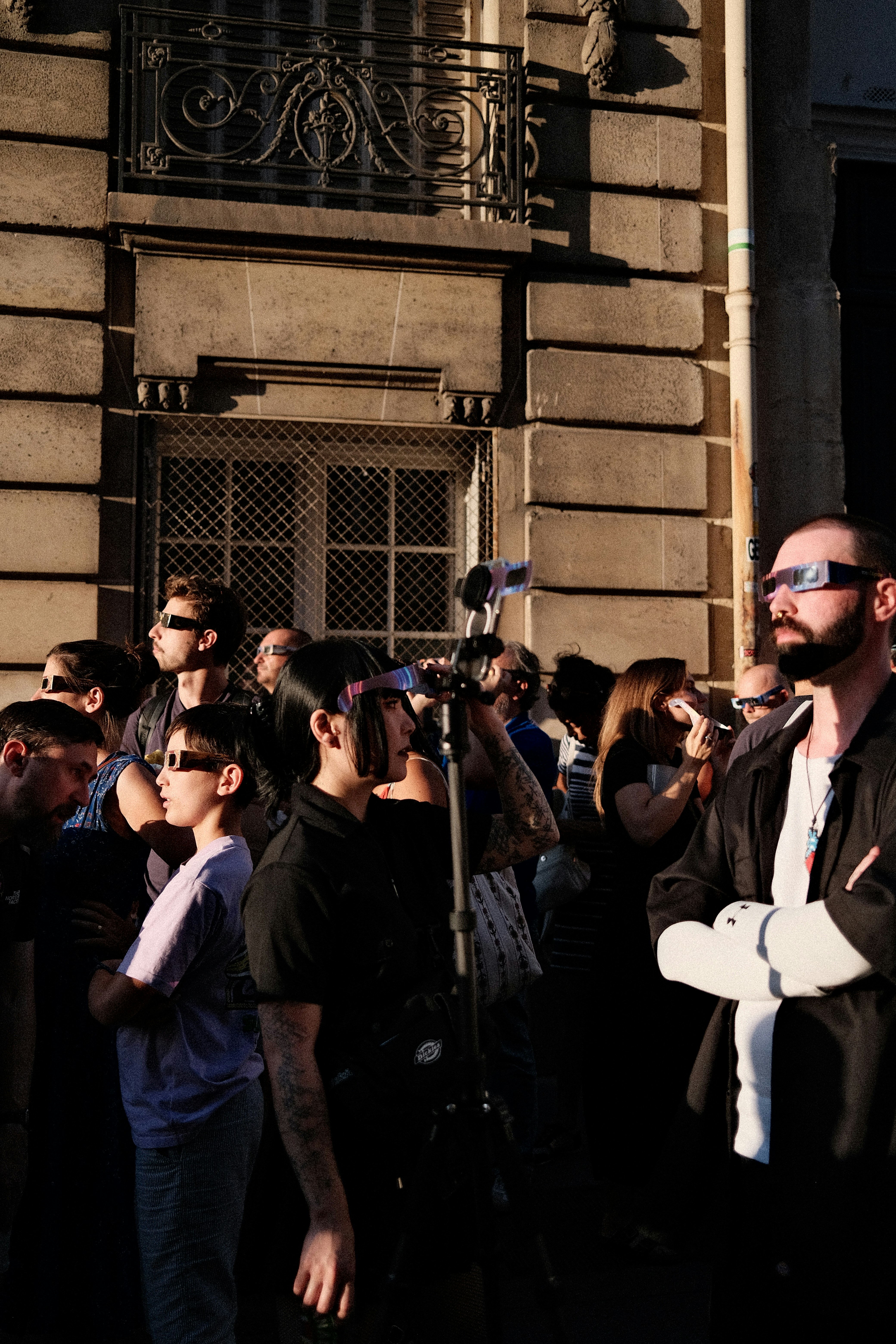 A group of people wearing solar eclipse glasses on a sunlit city street.