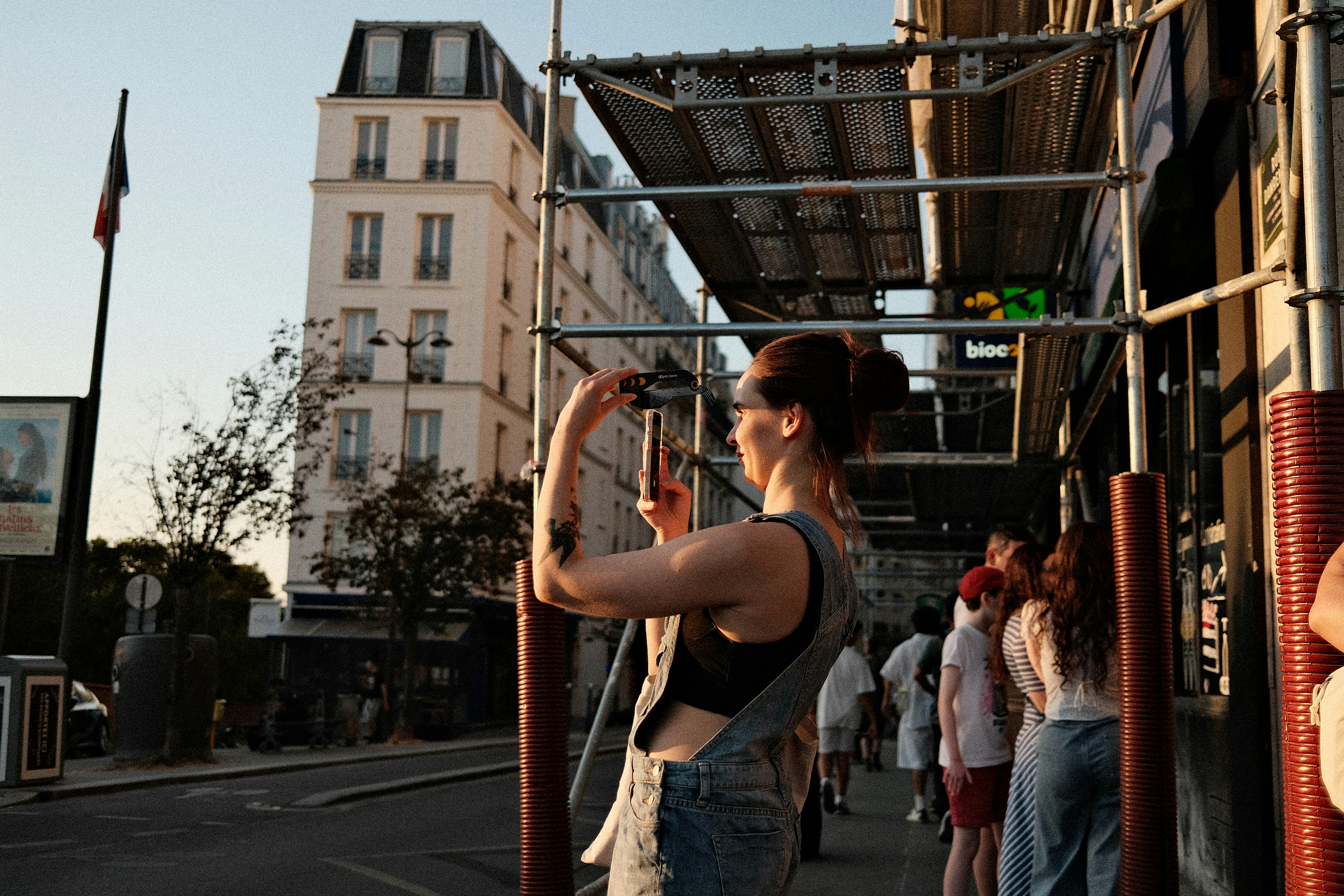 A woman in denim overalls photographing a street scene in Paris at golden hour