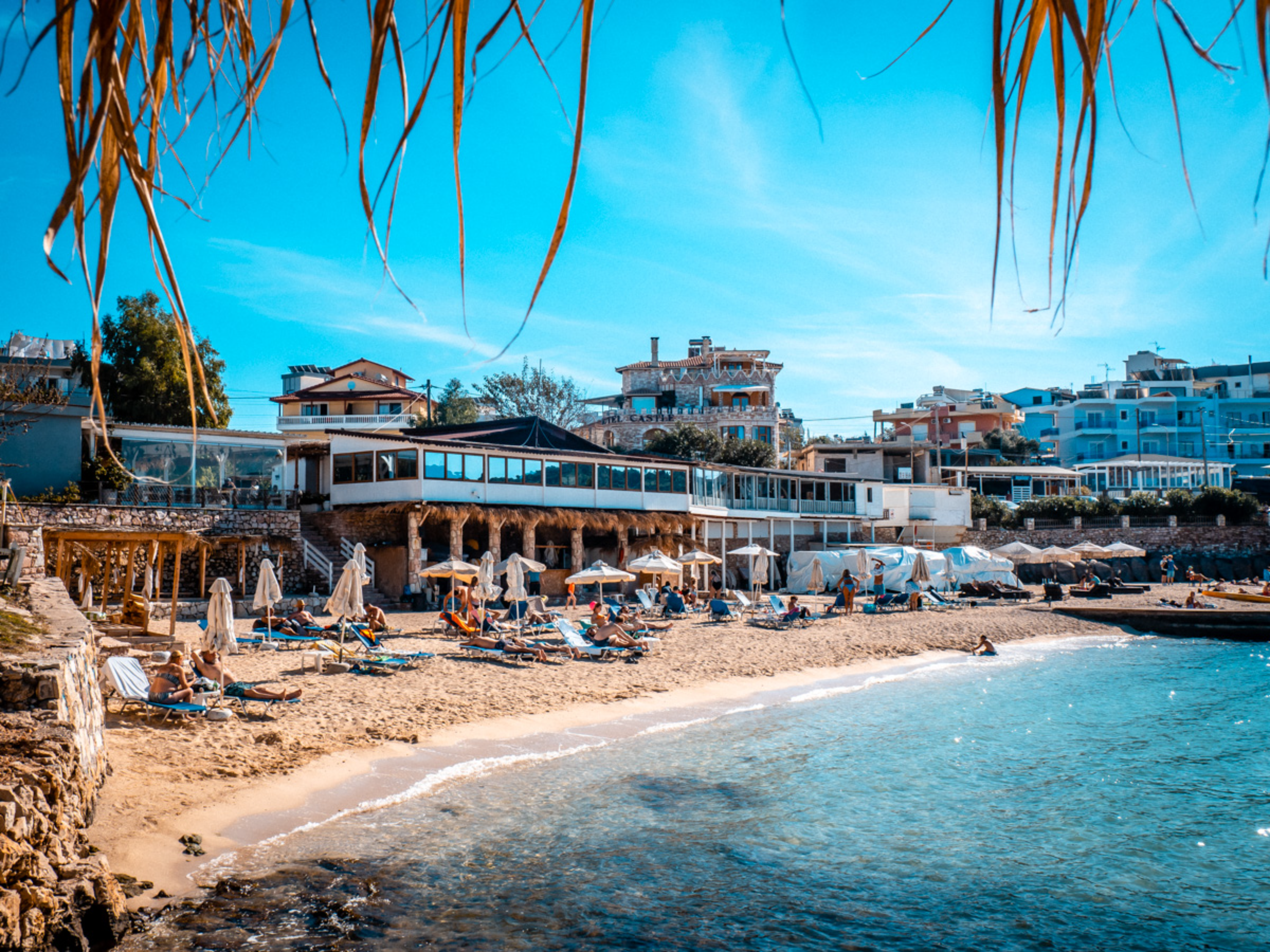People relaxing on lounge chairs on a sandy beach below a seaside restaurant