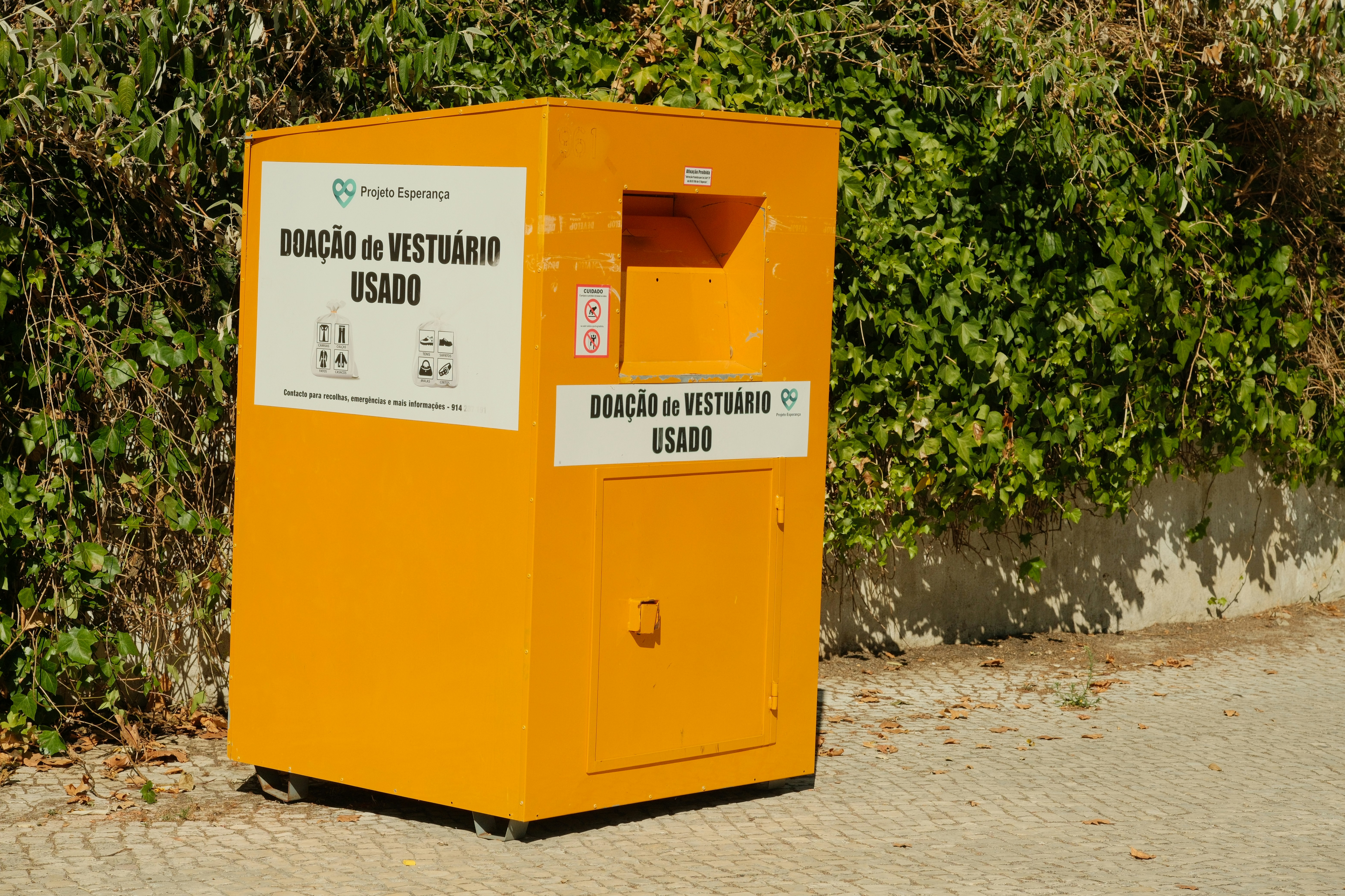 A bright yellow donation bin for used clothing against a green ivy wall