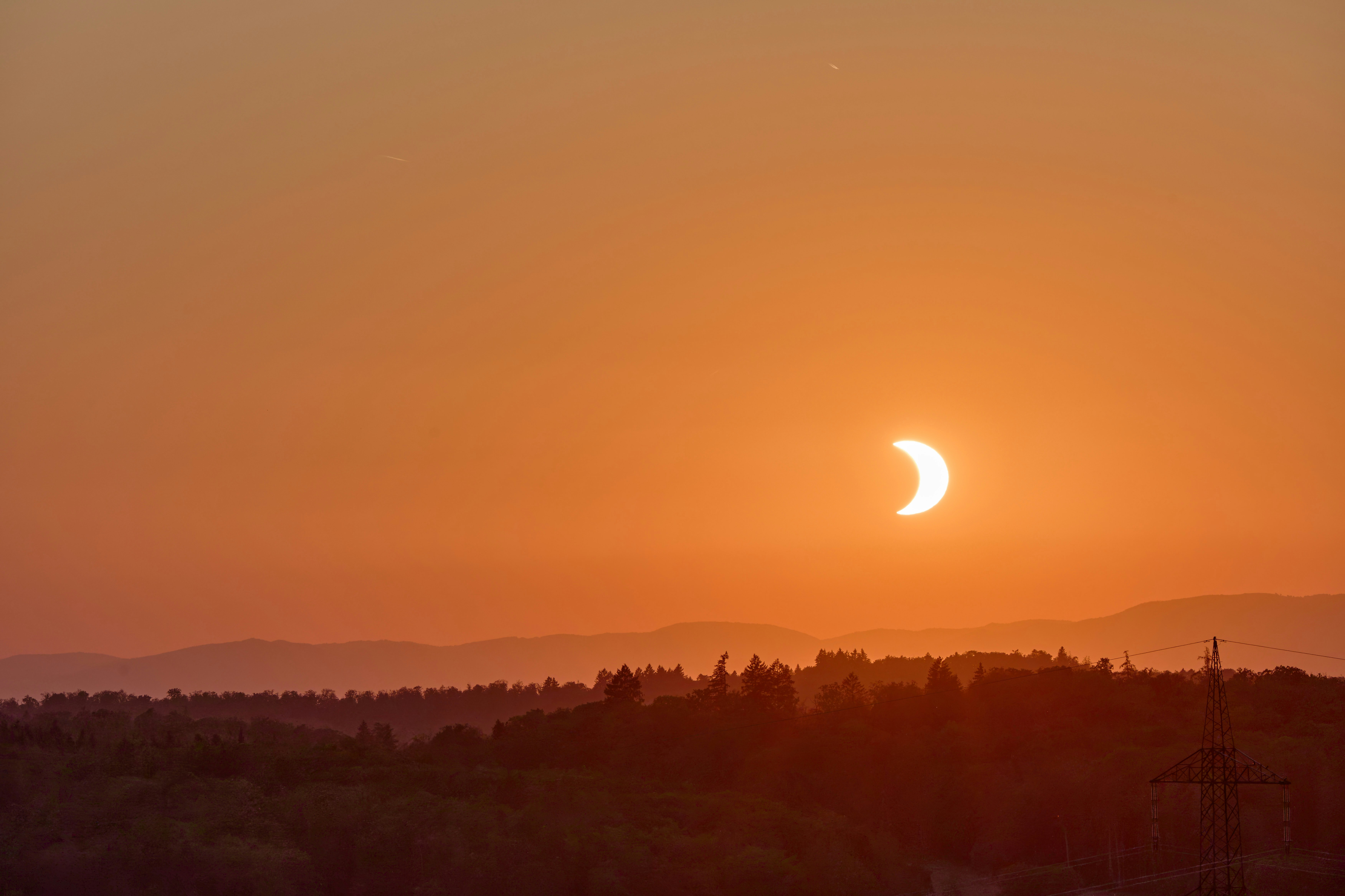 A partial solar eclipse over a dark forest and distant hills at golden hour.