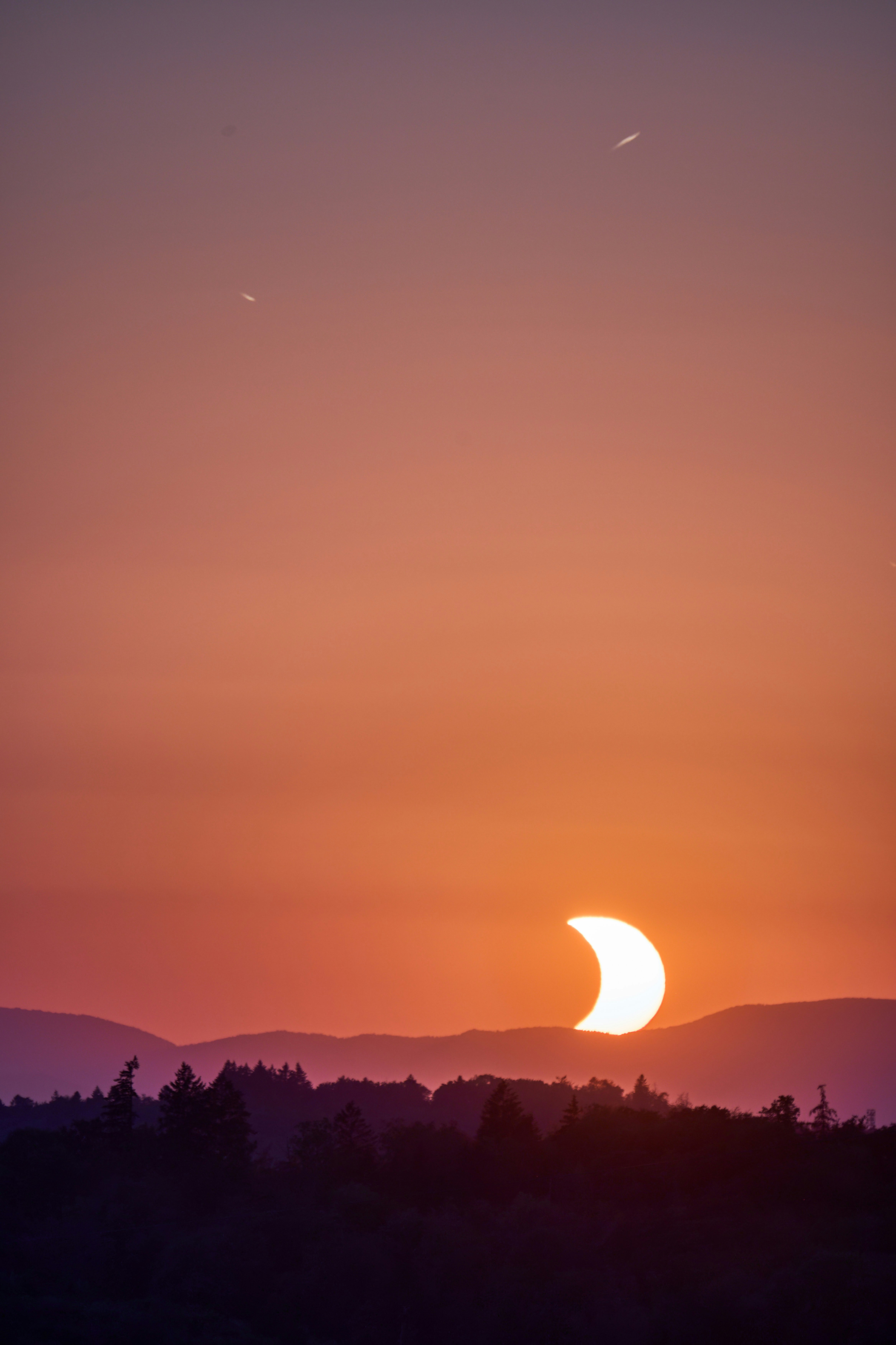 A partial solar eclipse above a dark mountain silhouette in a warm orange sky.