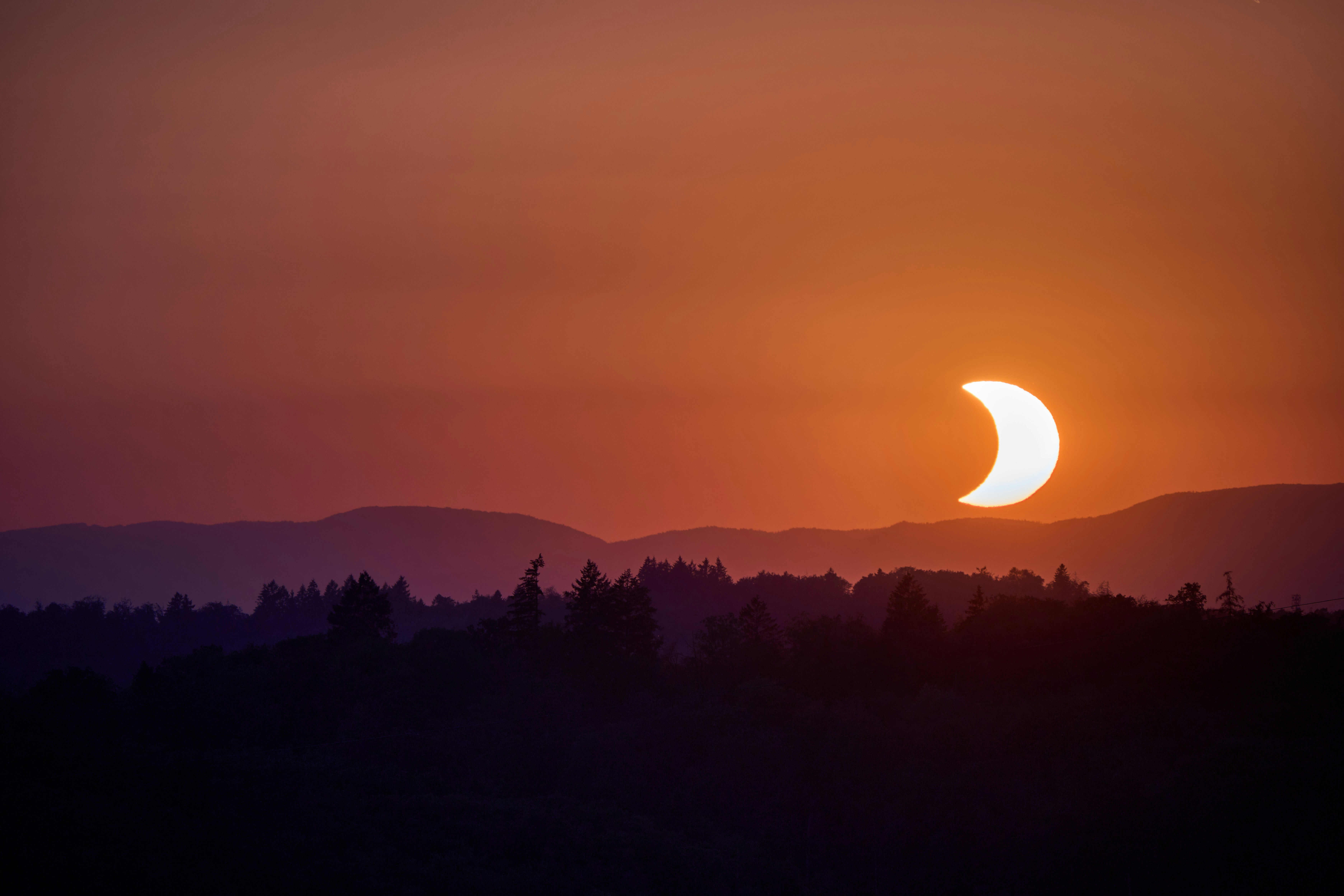A partial solar eclipse above a dark forest and distant mountain range.