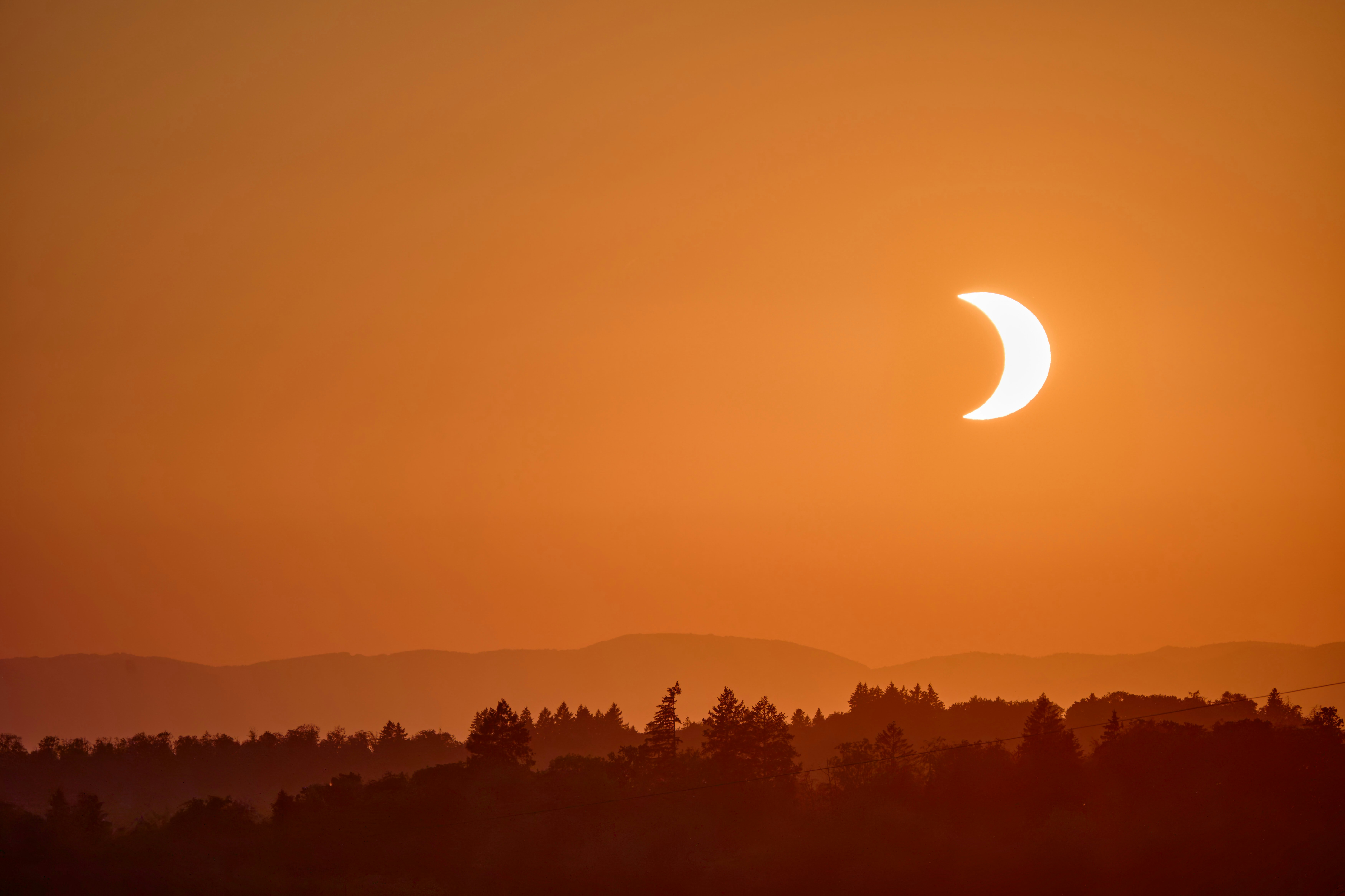 A crescent sun during a solar eclipse above a dark forest silhouette.