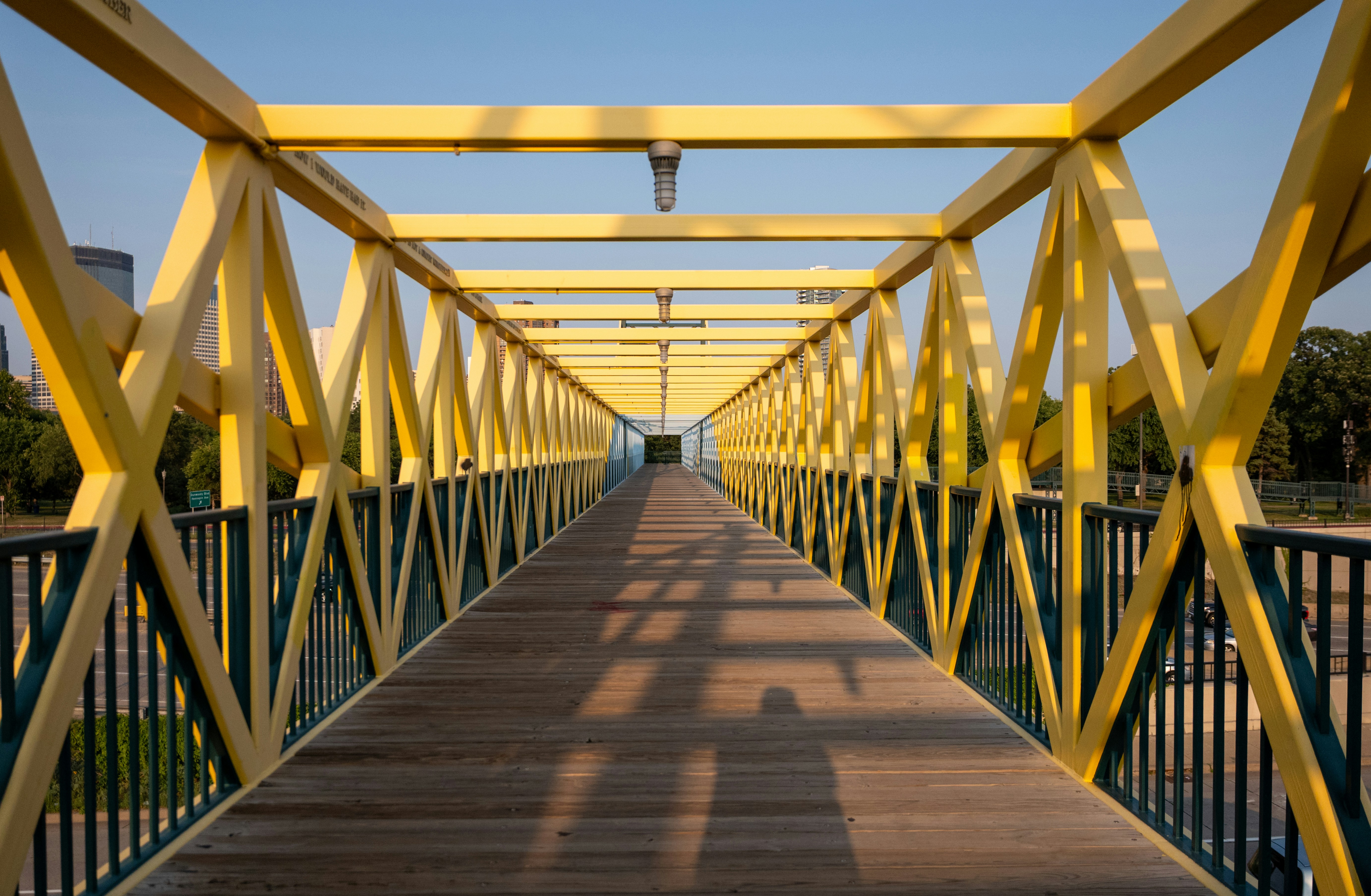 A pedestrian bridge with yellow steel trusses and a wooden walkway at low sun