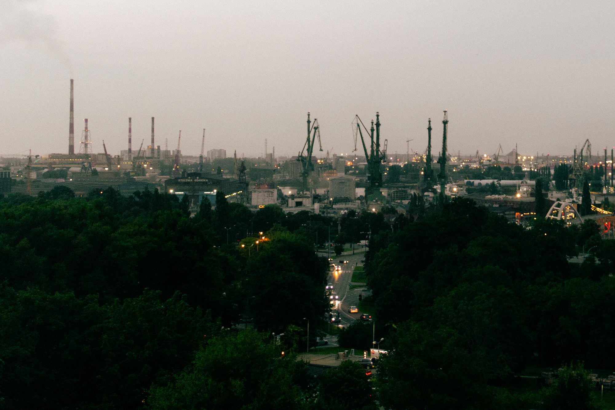 Industrial shipyard with tall cranes and stacks behind a dark treeline
