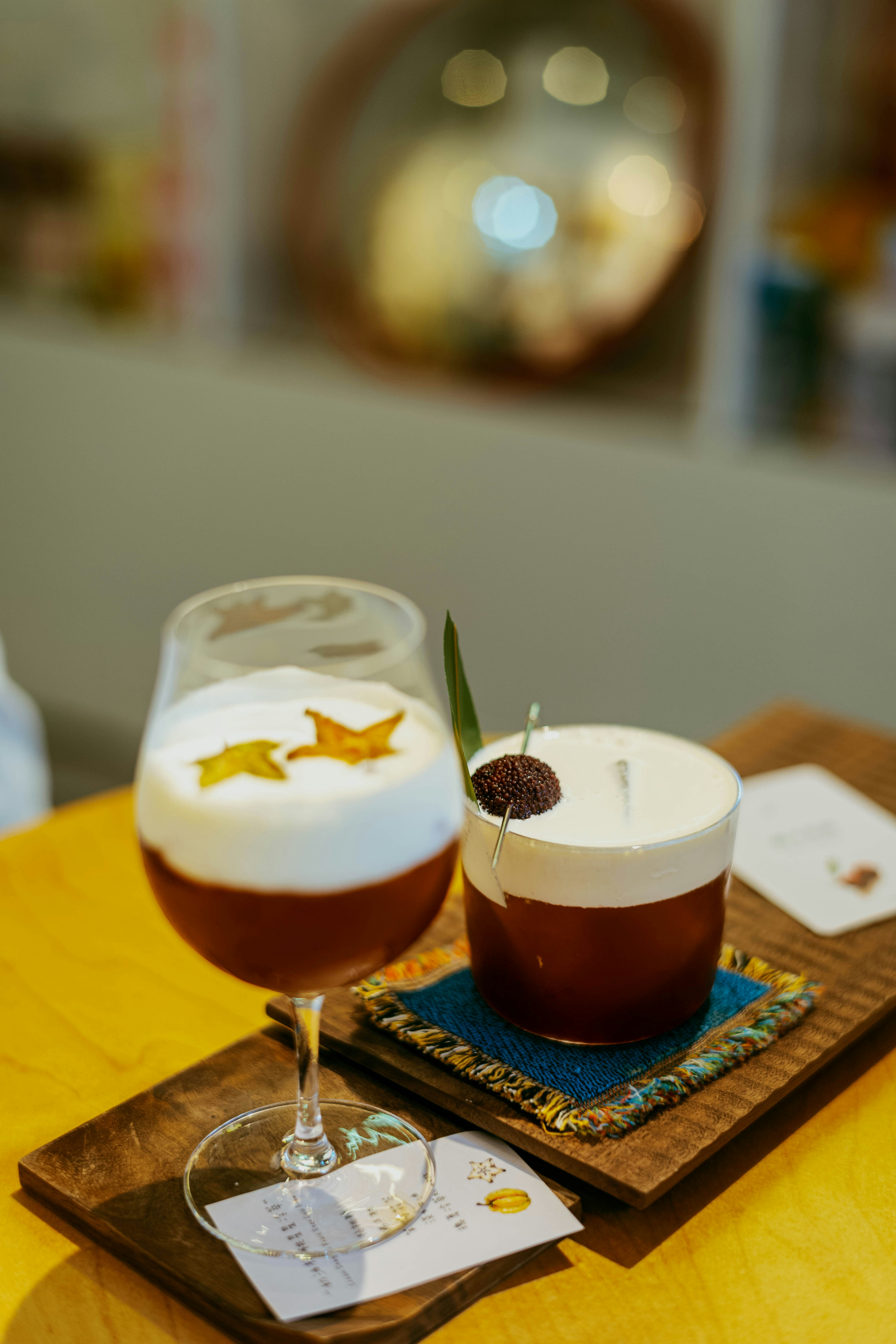 Two cocktails with foam toppings on a wooden tray in a cafe.