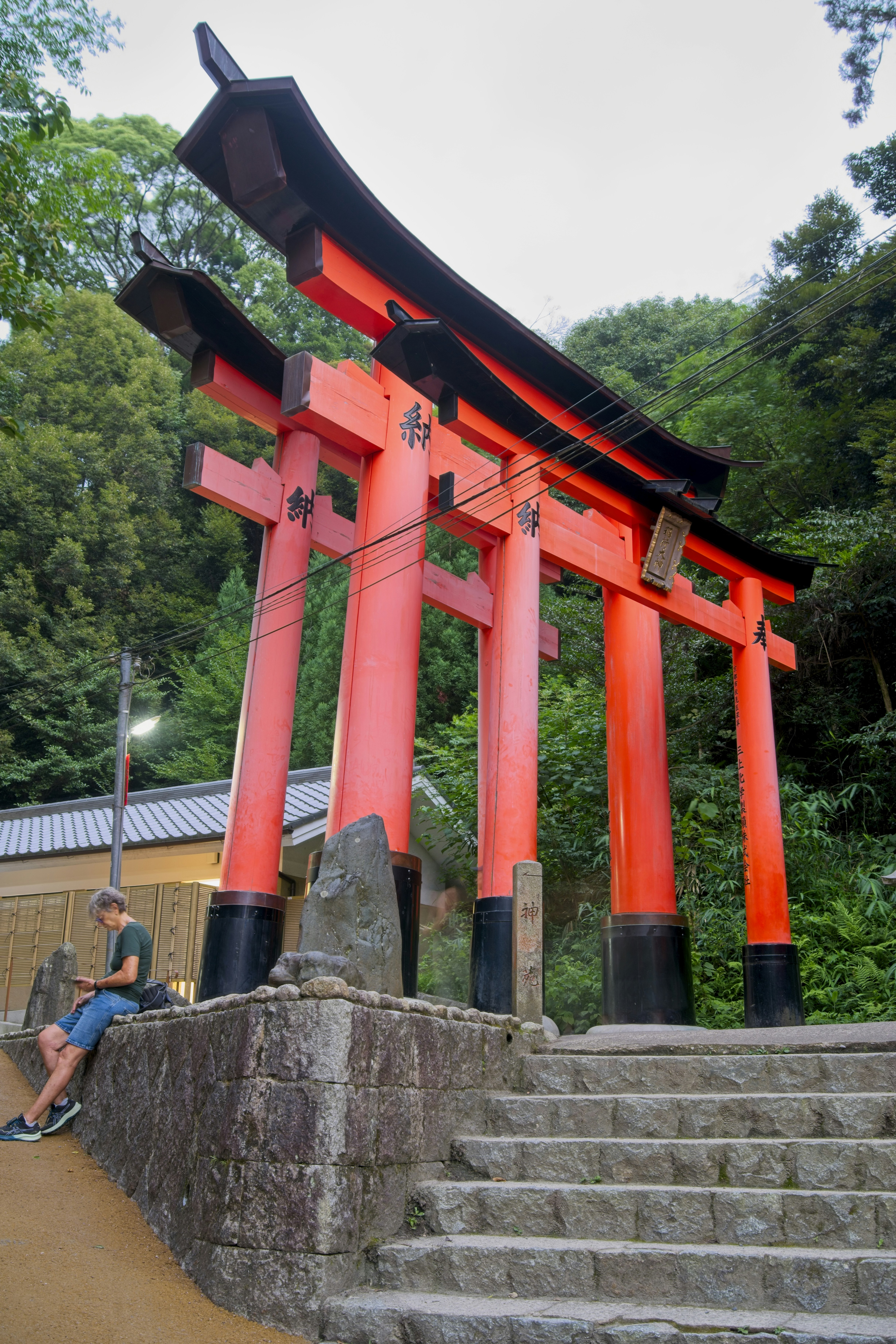 Una persona sentada en un muro de piedra junto a una gran puerta torii roja.