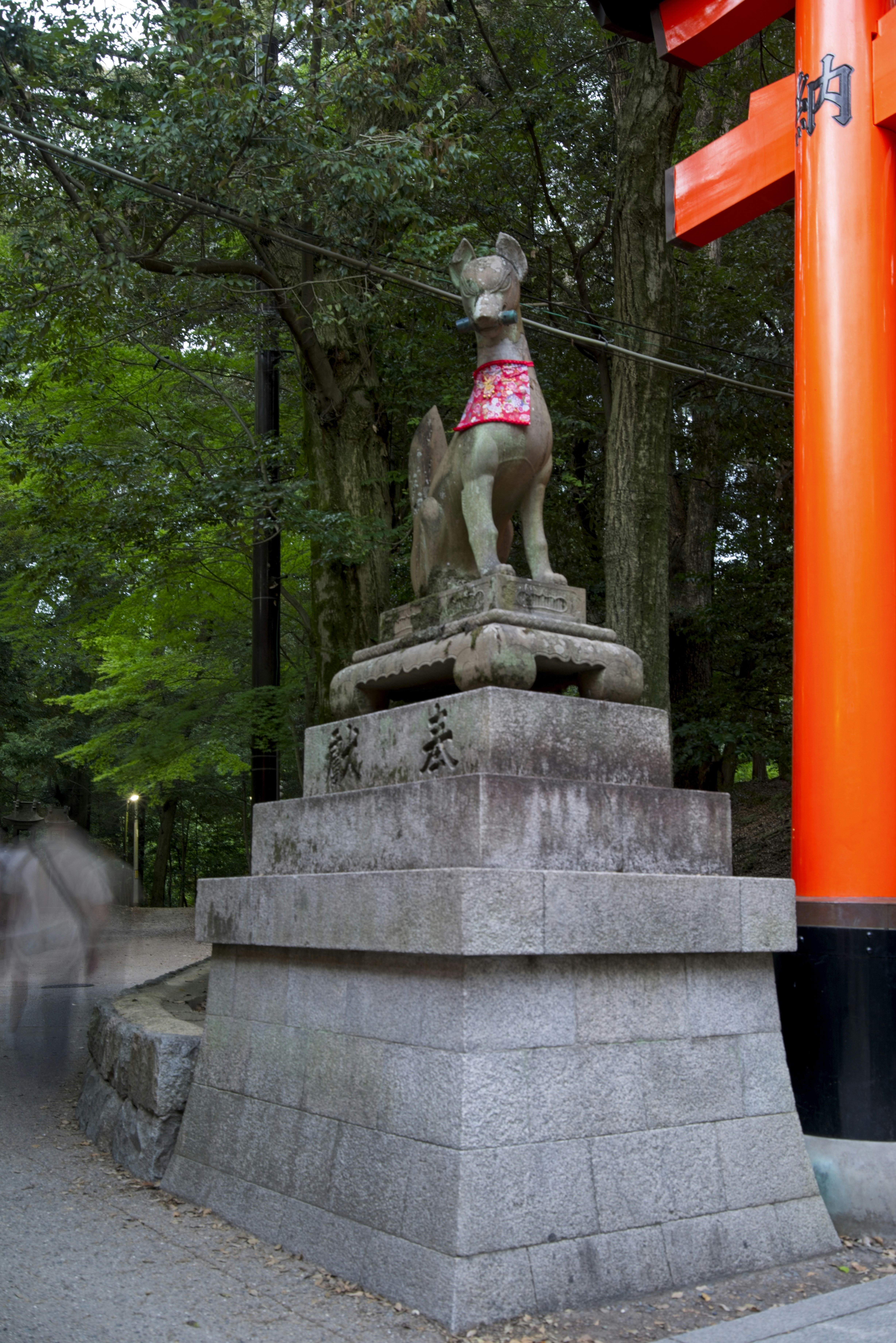 Una estatua de zorro de piedra con un babero rojo con estampado junto a una puerta torii naranja brillante.