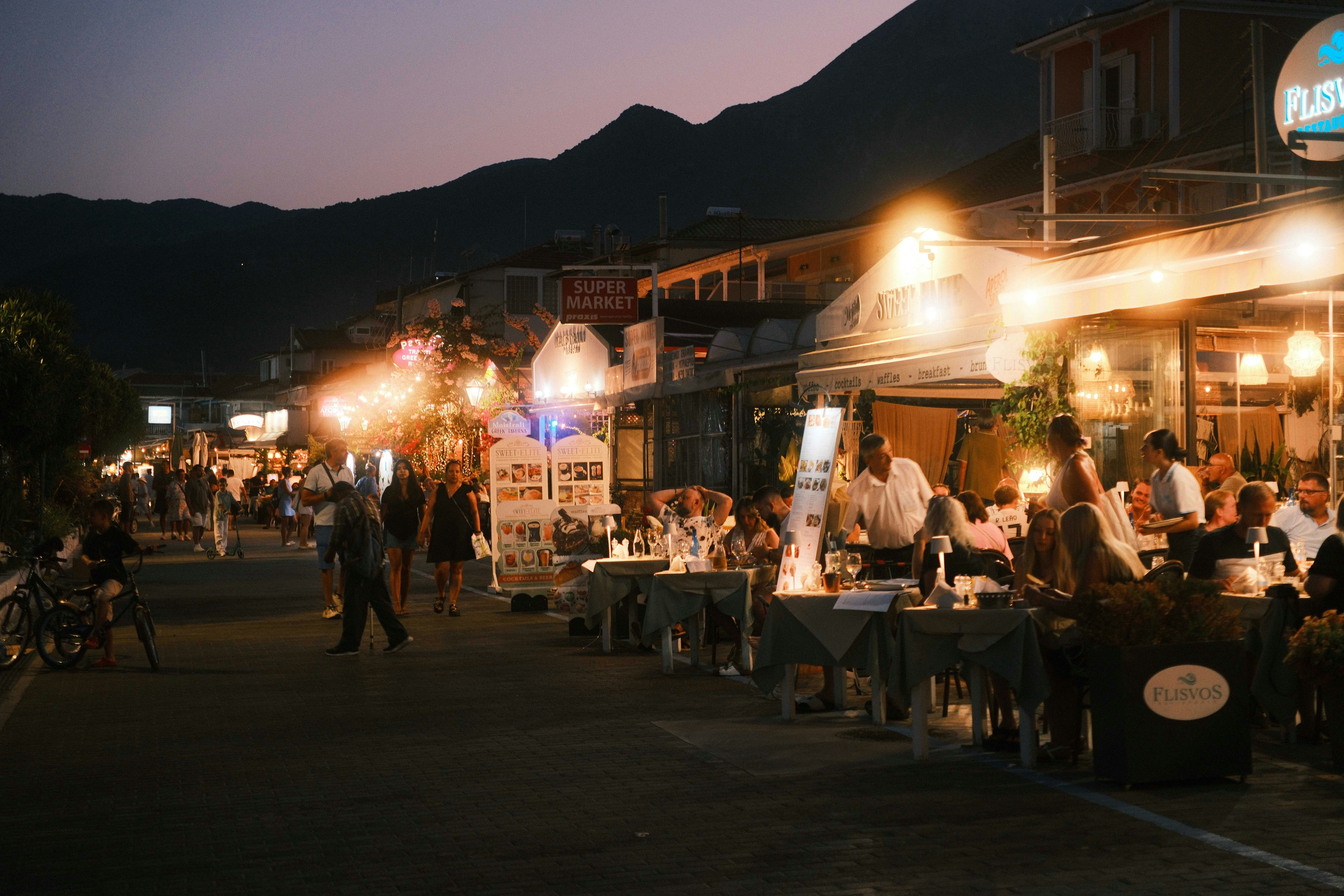 A village street at night with people eating at long tables outside lit shops, mountains in the dark behind