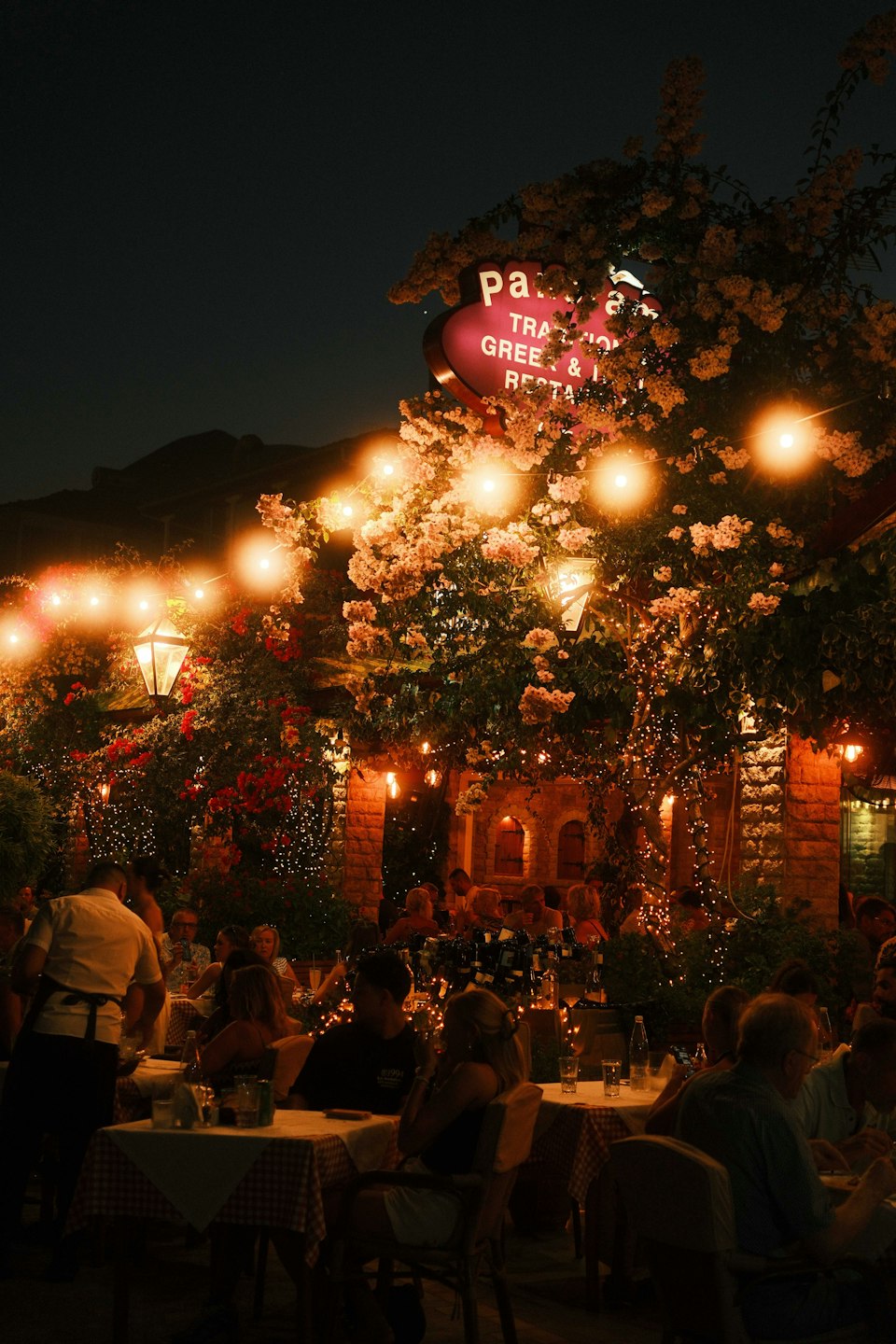 A Greek taverna terrace strung with lights and packed with diners at night