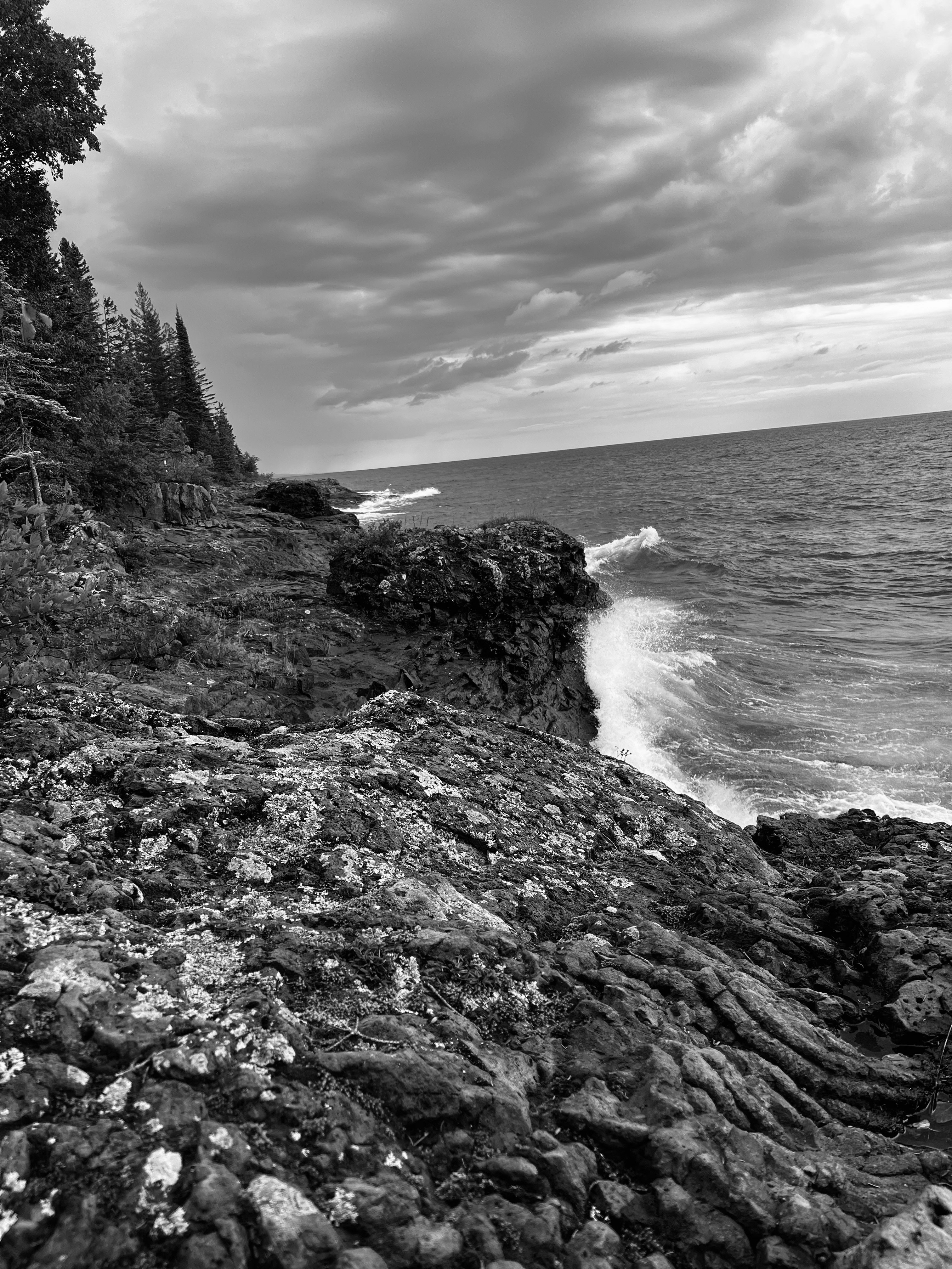Waves crashing against a rocky shoreline under a cloudy sky in black and white