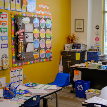 A colorful elementary school classroom with yellow walls and educational posters on a bulletin board