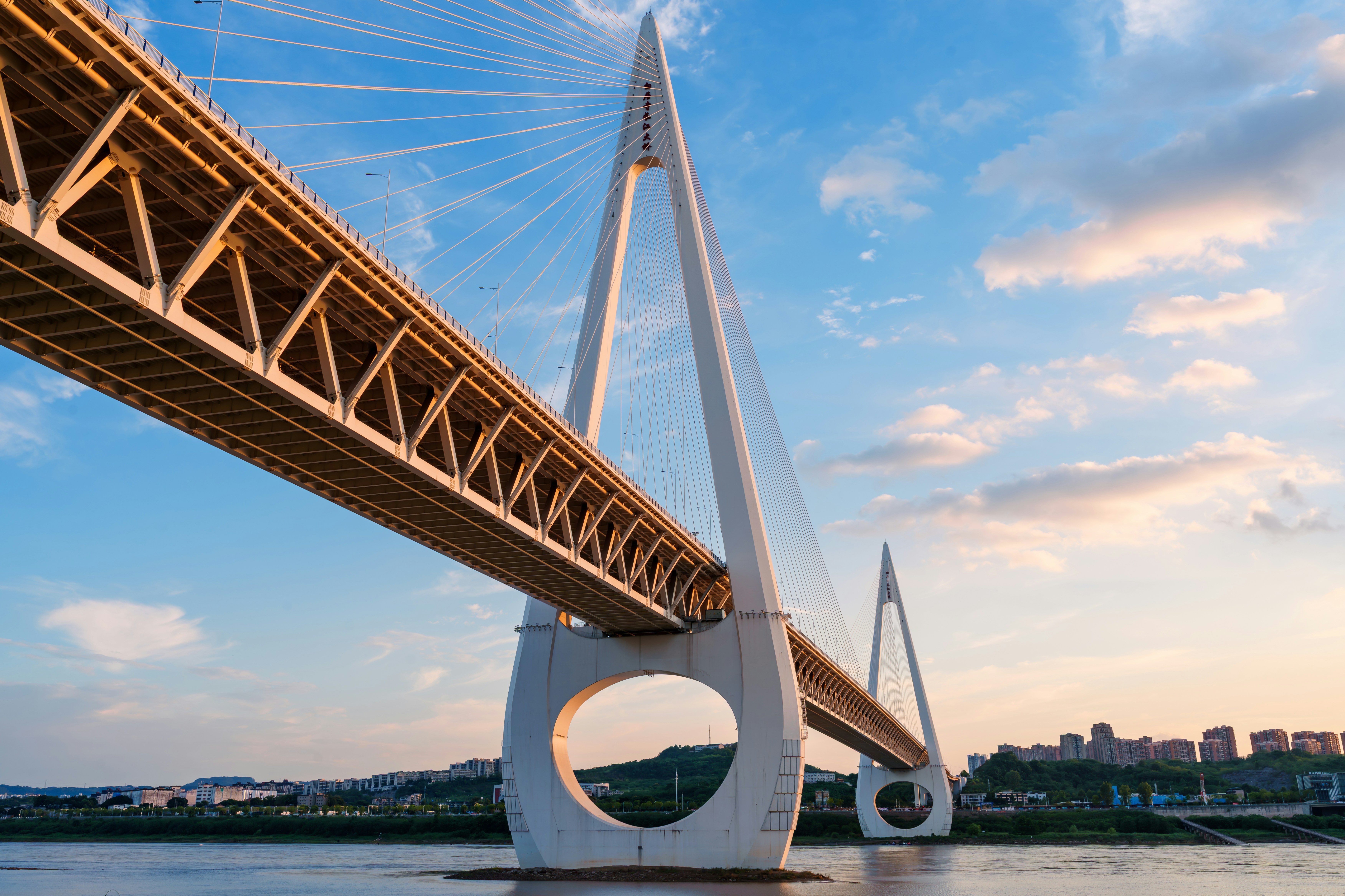 A white cable-stayed bridge with circular pylon bases over a river at golden hour