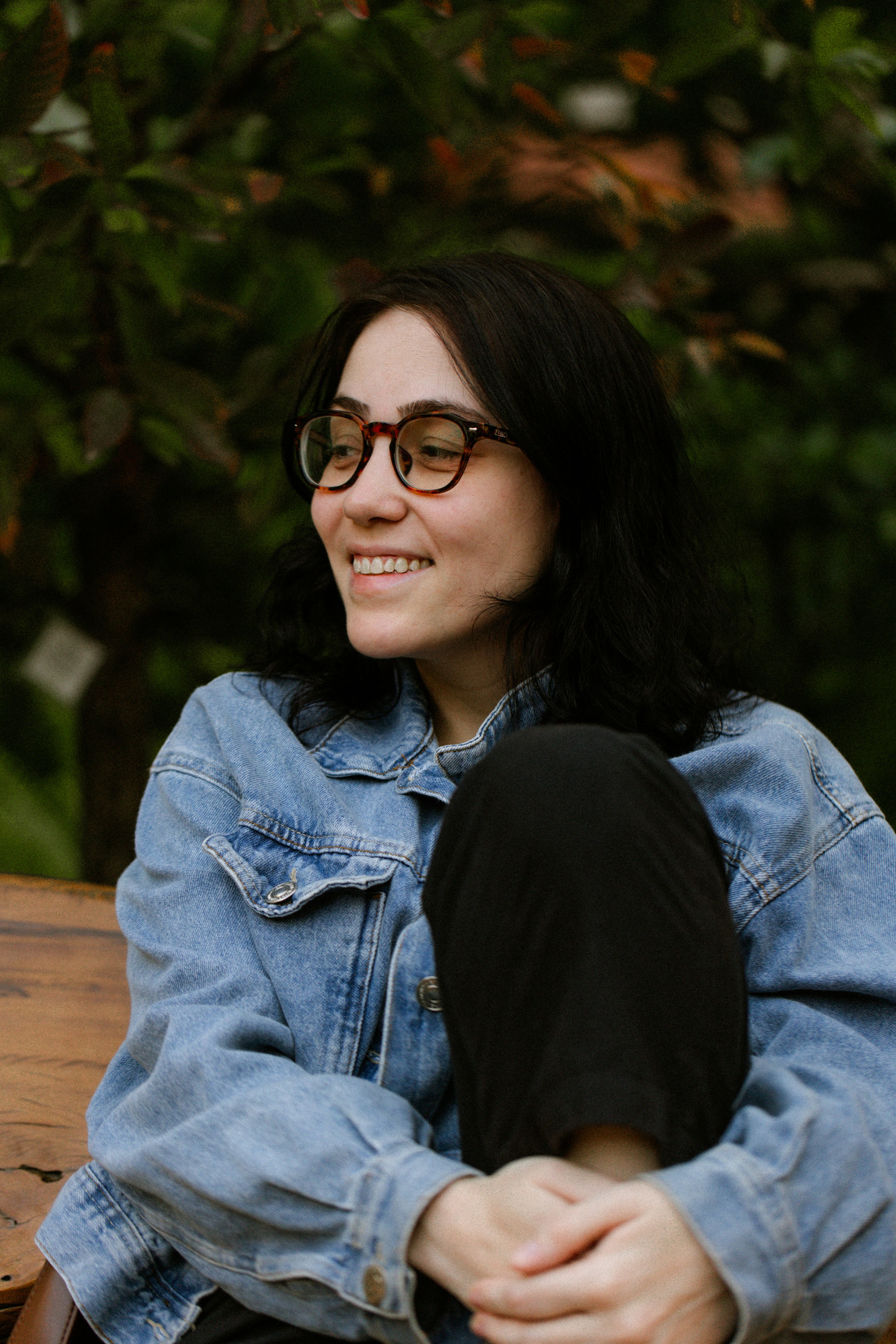 A woman with dark hair and glasses wearing a denim jacket outdoors.